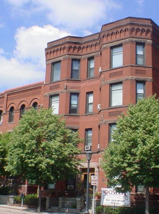 Aeon Affordable Homes and Community Affordable apartment homes in a red brick building with arched windows, nestled among trees, against a backdrop of a tall modern structure.