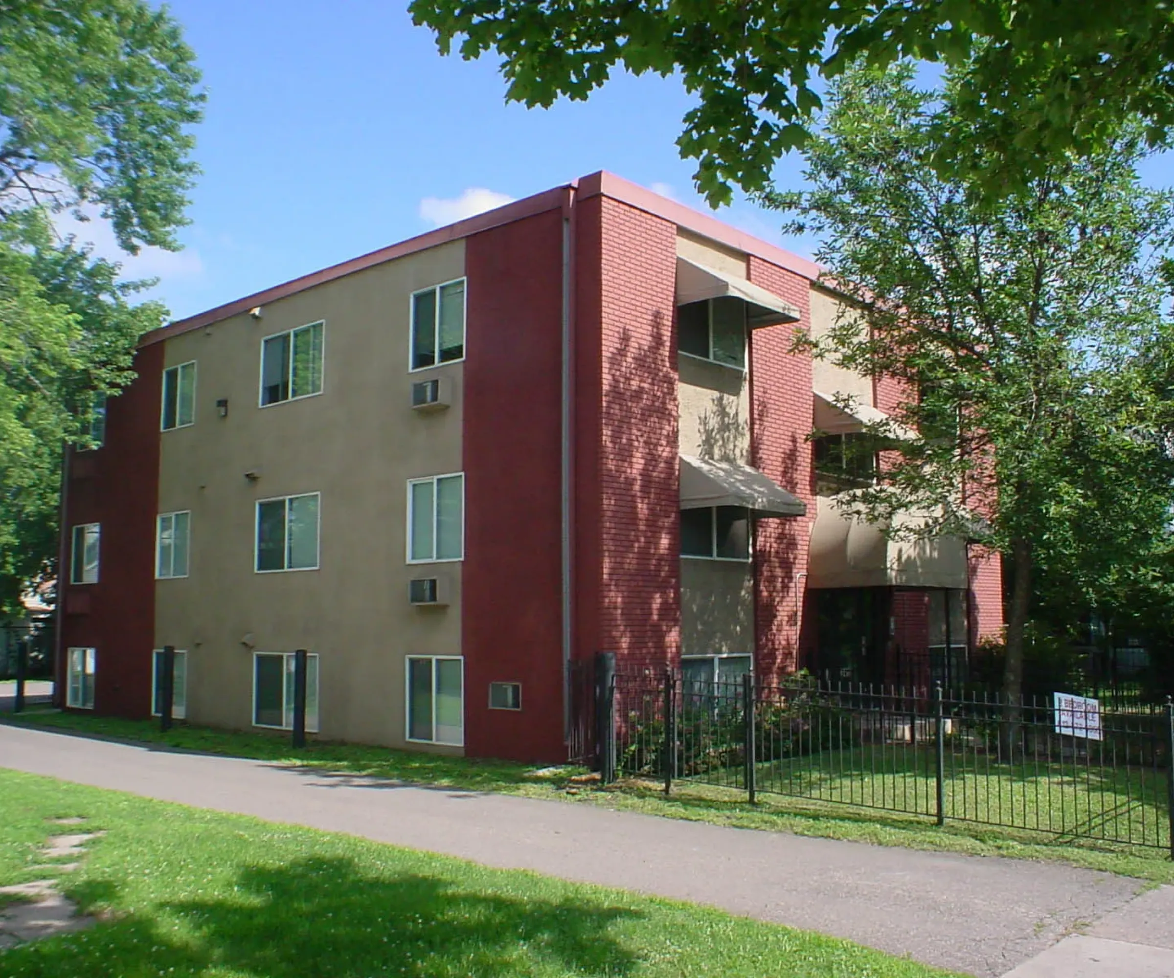 Aeon Affordable Homes and Community Affordable apartment homes with a tan and red exterior, surrounded by trees and a black fence, under a clear blue sky.