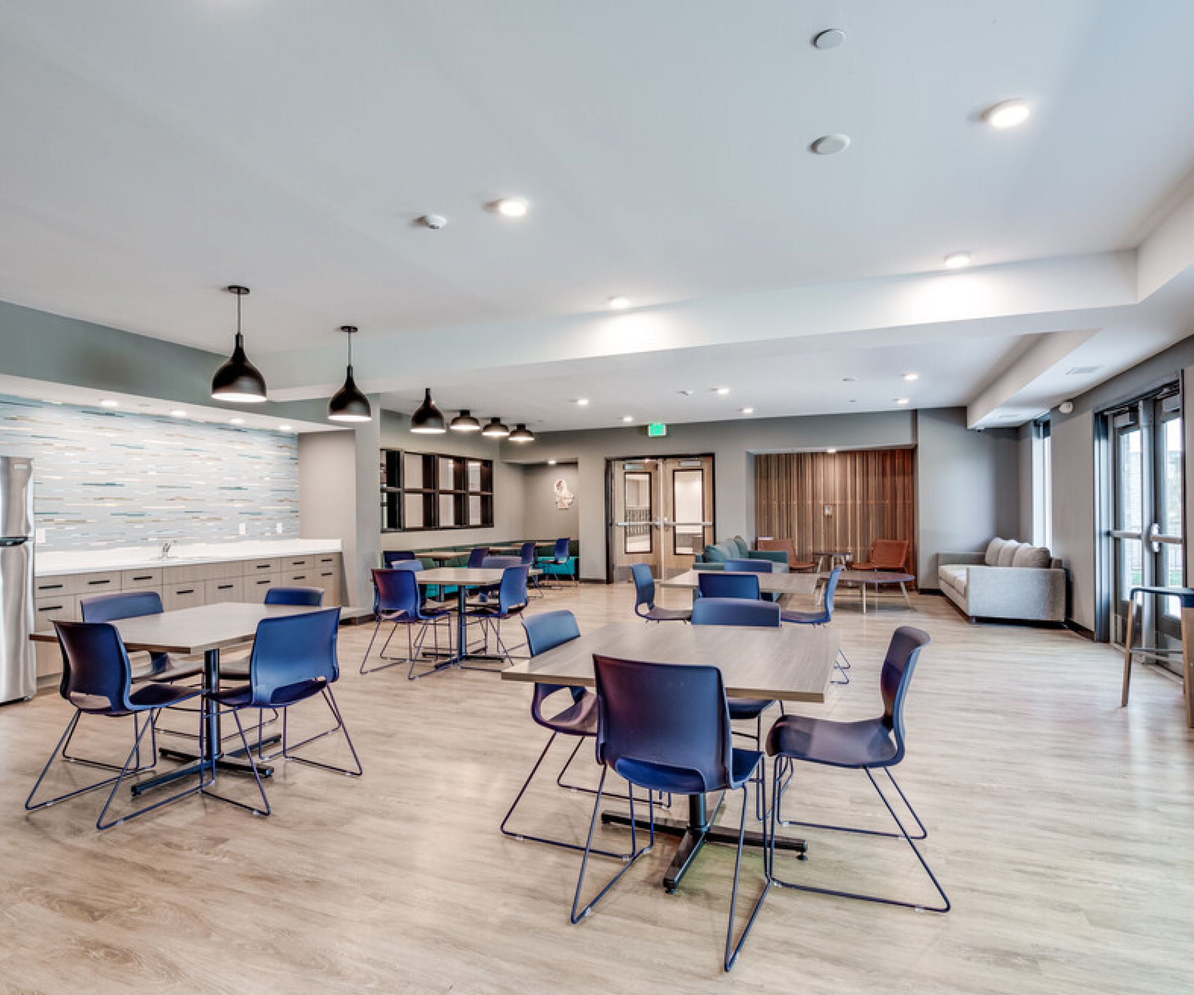 Aeon Affordable Homes and Community Affordable apartment homes featuring a modern communal room with blue chairs, tables, kitchen area, pendant lights, and wood flooring.