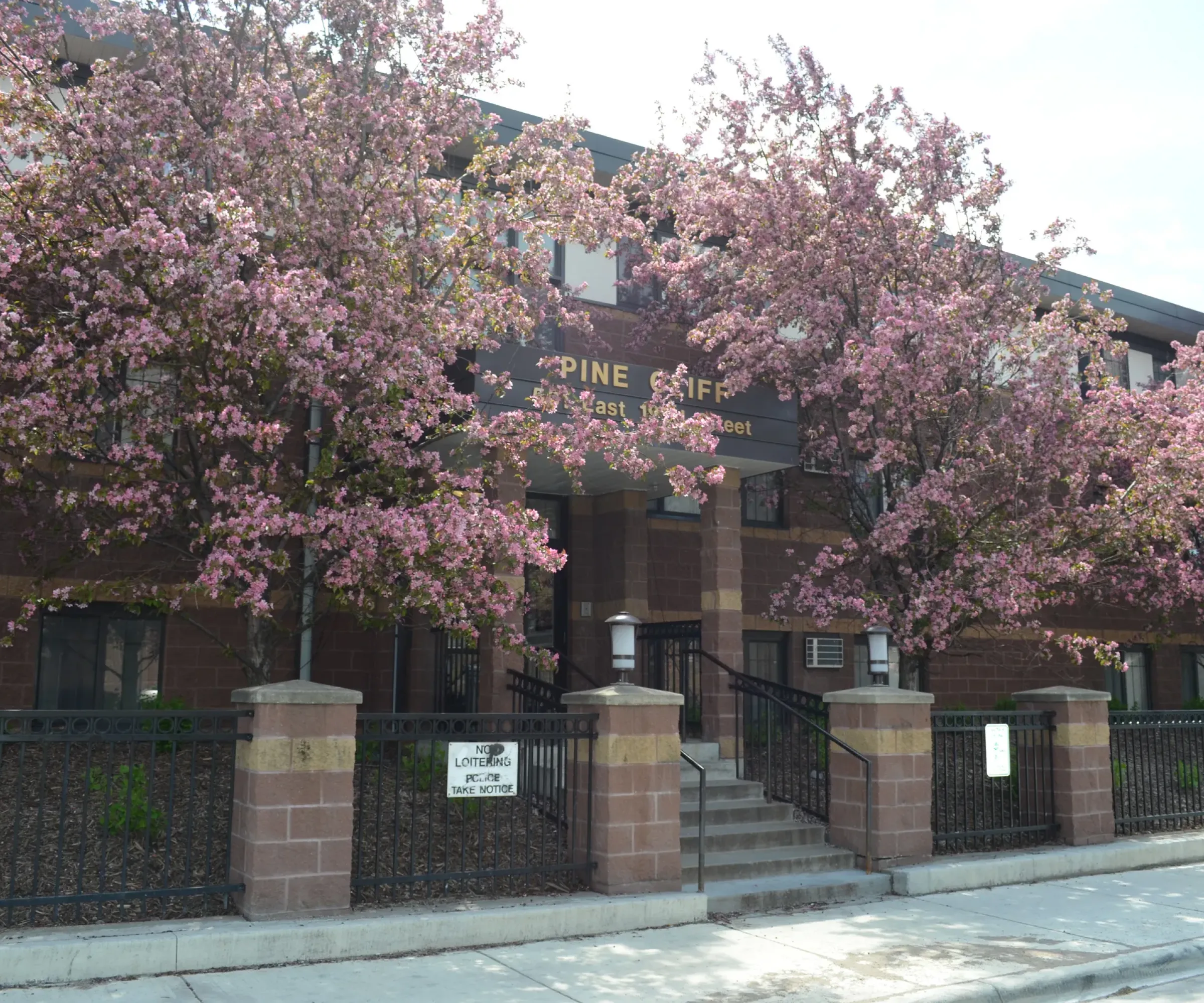 Aeon Affordable Homes and Community A brick apartment building with blooming pink trees, a black fence, and affordable apartment homes awaits in front.