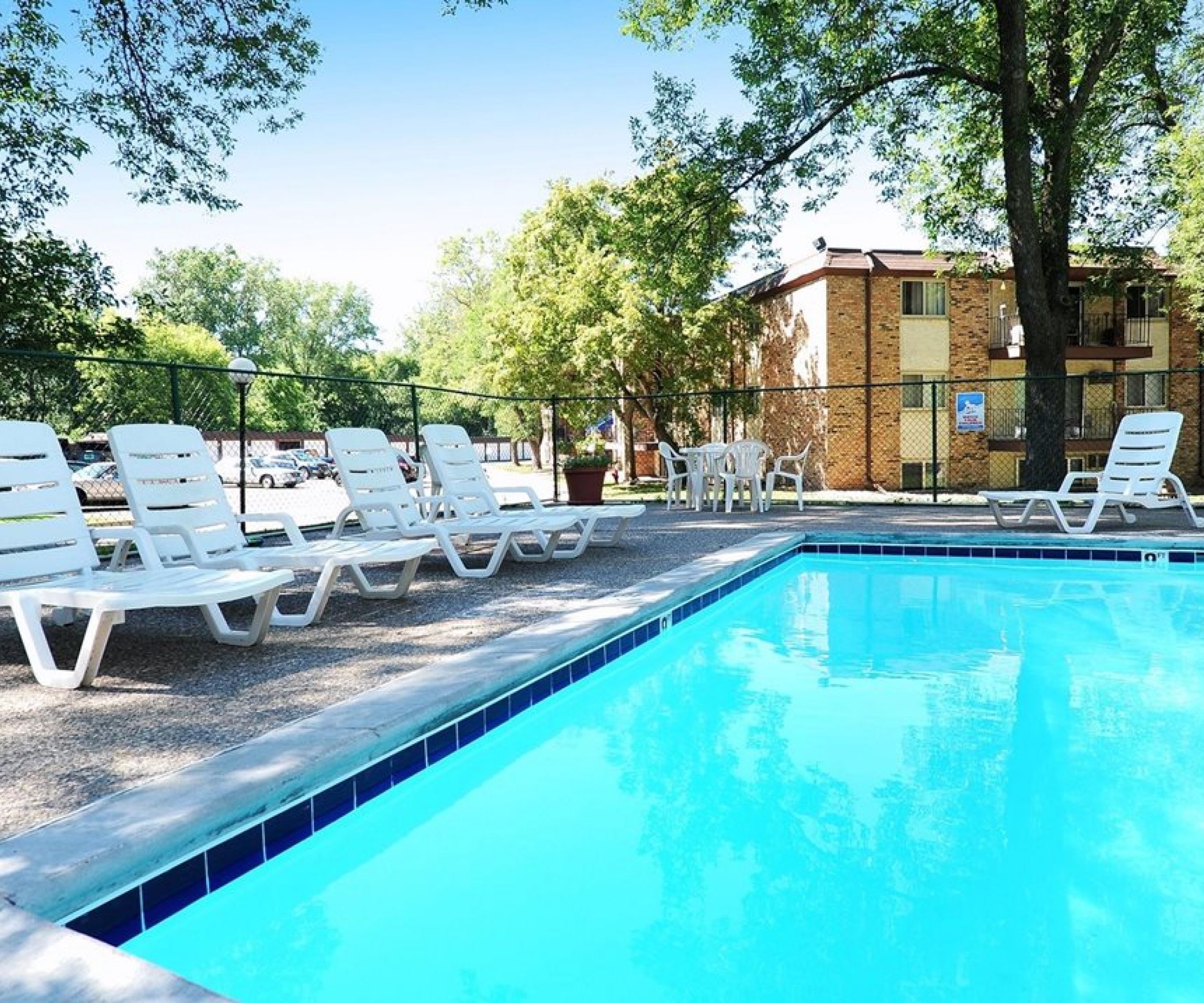 Aeon Affordable Homes and Community Affordable apartment homes with an outdoor pool feature blue water, white lounge chairs, and trees under a clear sky.