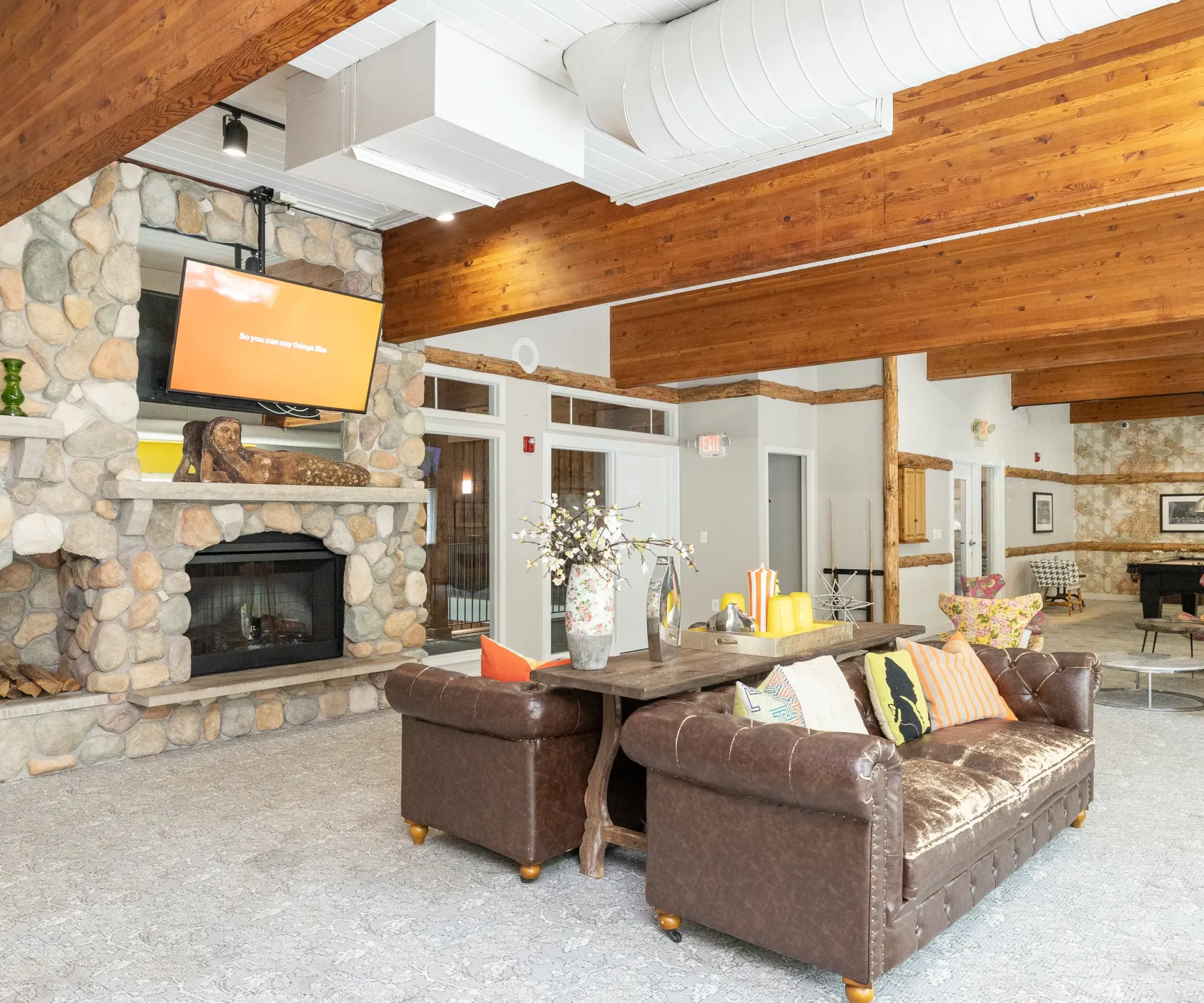 Aeon Affordable Homes Lounge with stone fireplace, TV, leather couch, and wooden beams in Blooming Meadows South; decorated with vases and cushions.