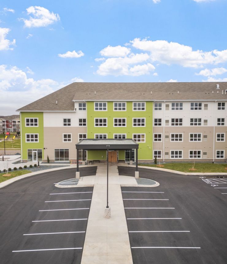 Aeon Affordable Homes and Community Large, four-story building with a green accent offering affordable apartment homes, surrounded by an empty parking lot and partly cloudy sky.