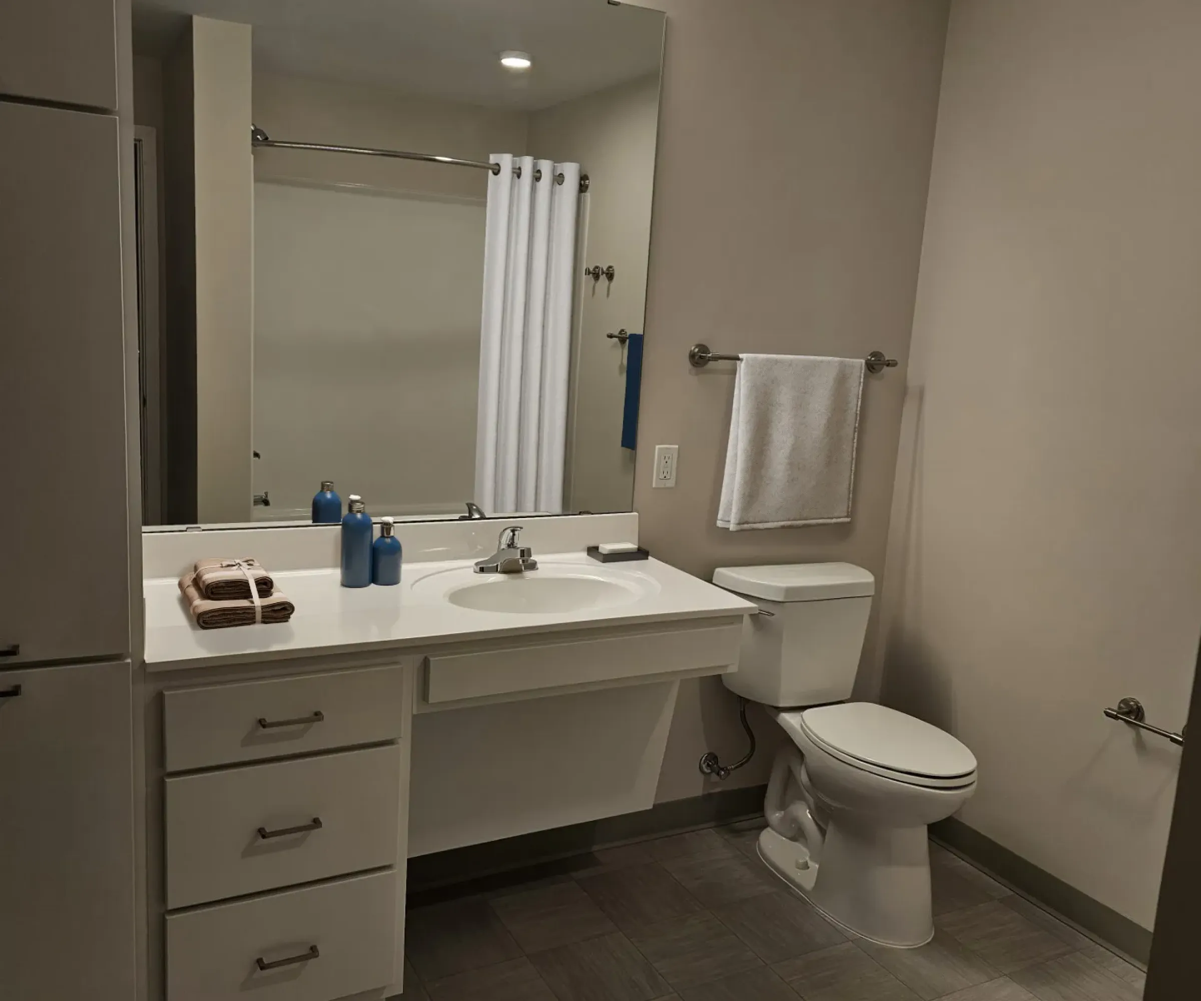 Aeon Affordable Homes and Community Modern bathroom in affordable apartment homes with a large mirror, vanity with toiletries, toilet, towel rail, and shower curtain.