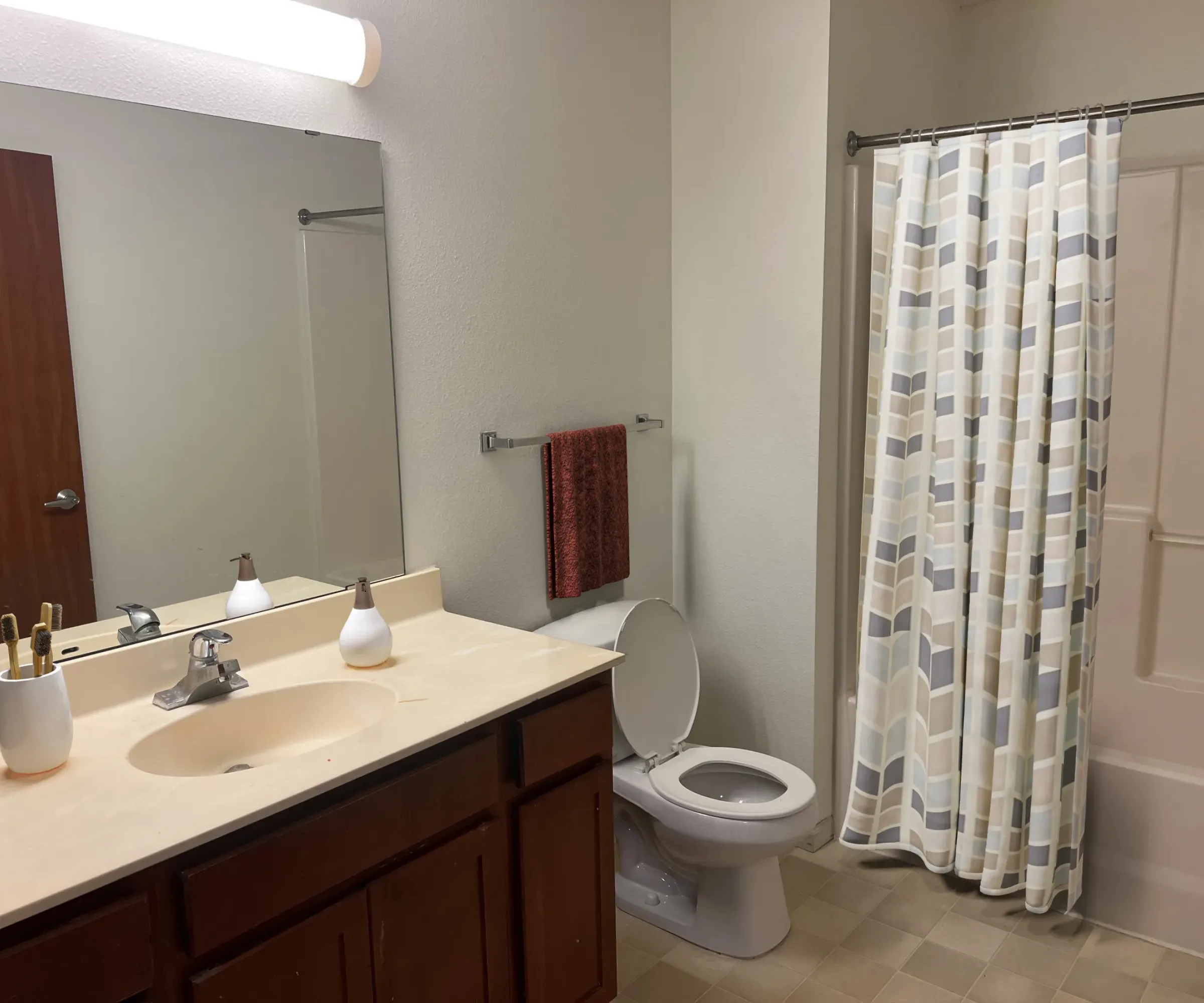 Aeon Affordable Homes and Community Affordable apartment homes feature a bathroom with a wooden vanity, large mirror, toilet, and bathtub with geometric-patterned shower curtain.