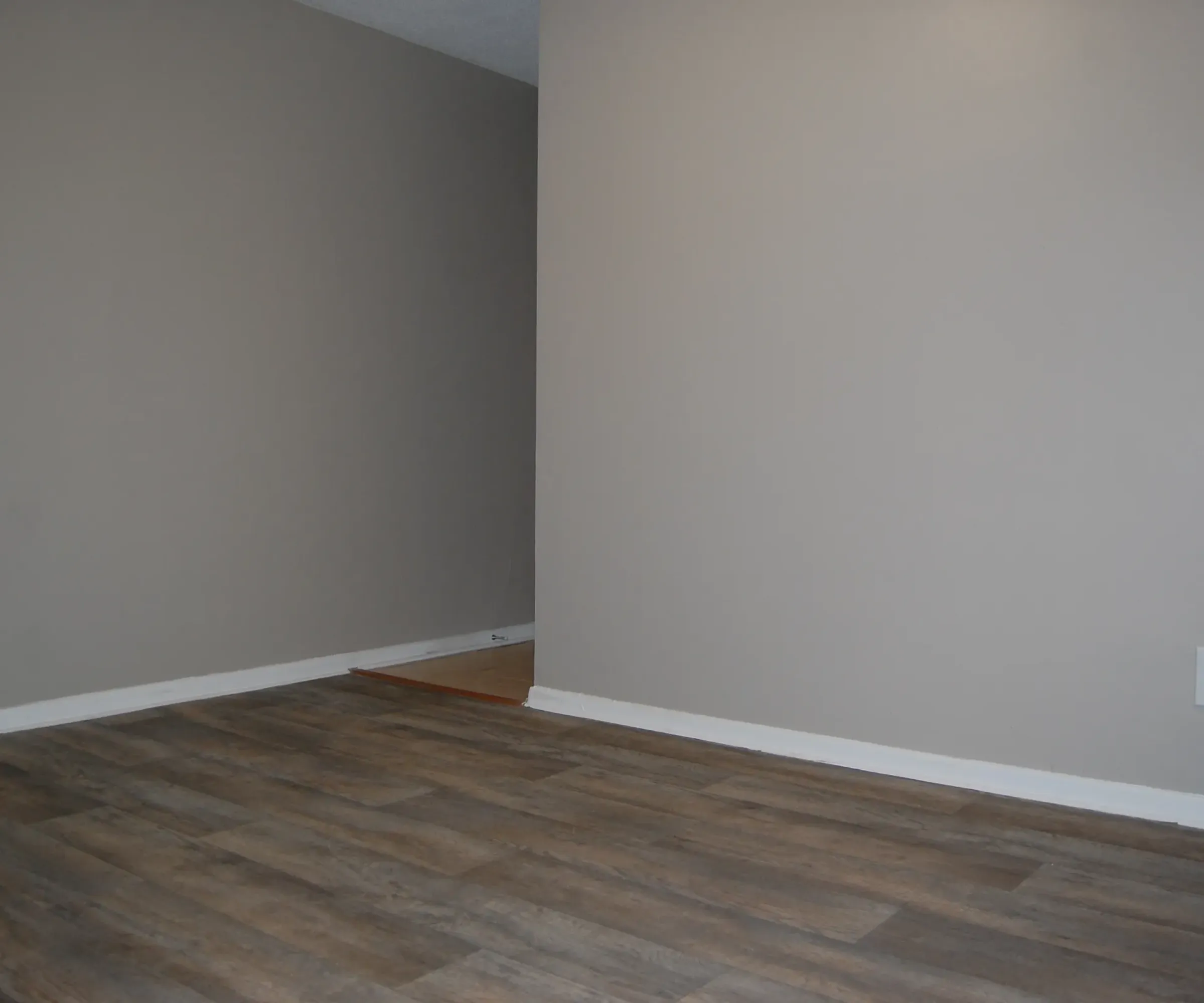 Aeon Affordable Homes and Community Affordable apartment homes feature an empty room with gray walls and a wooden floor, with a corner and small step leading to another area.