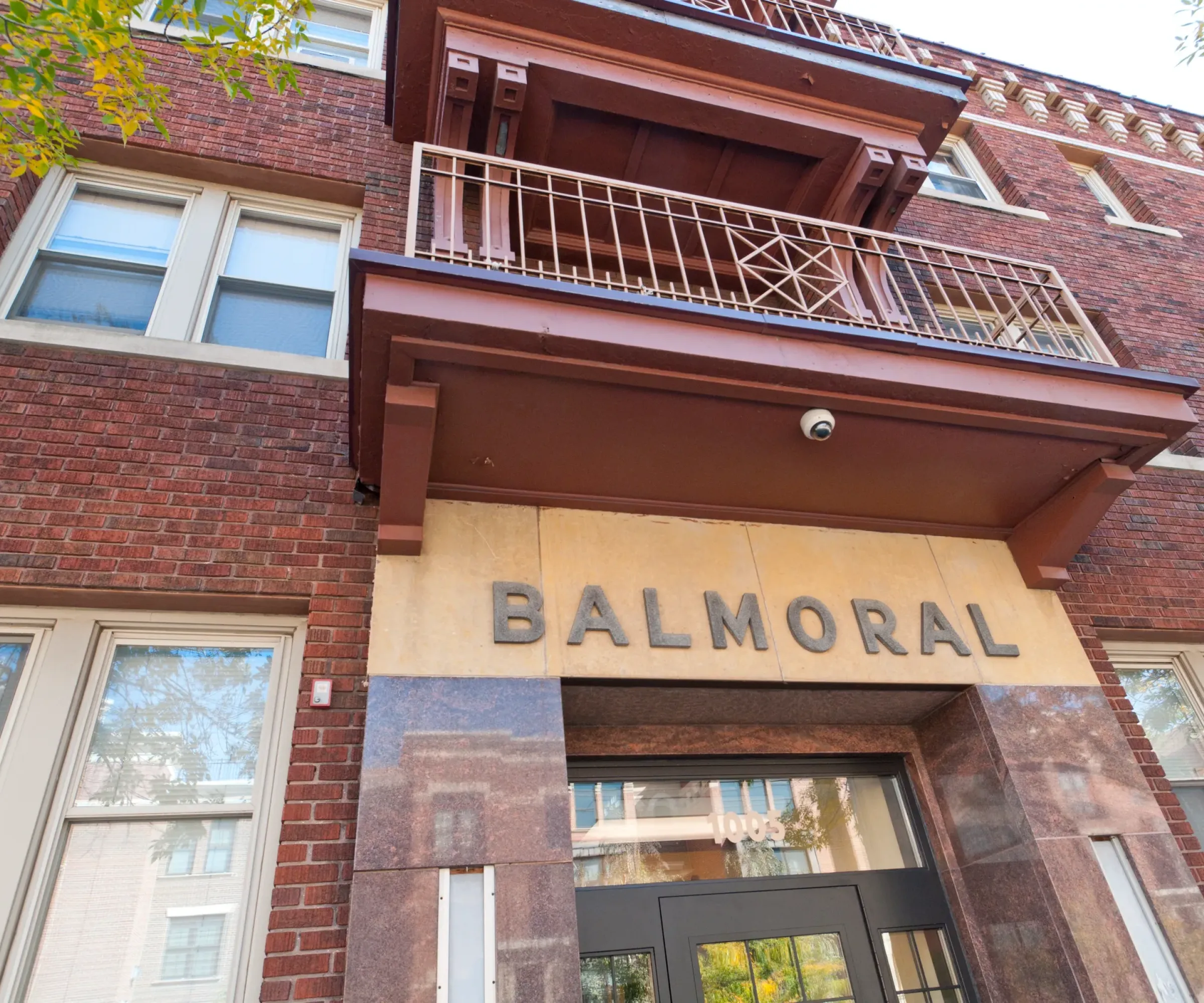 Aeon Affordable Homes and Community Brick building with "BALMORAL" over the entrance, featuring affordable apartment homes with large windows and a balcony with a metal railing.