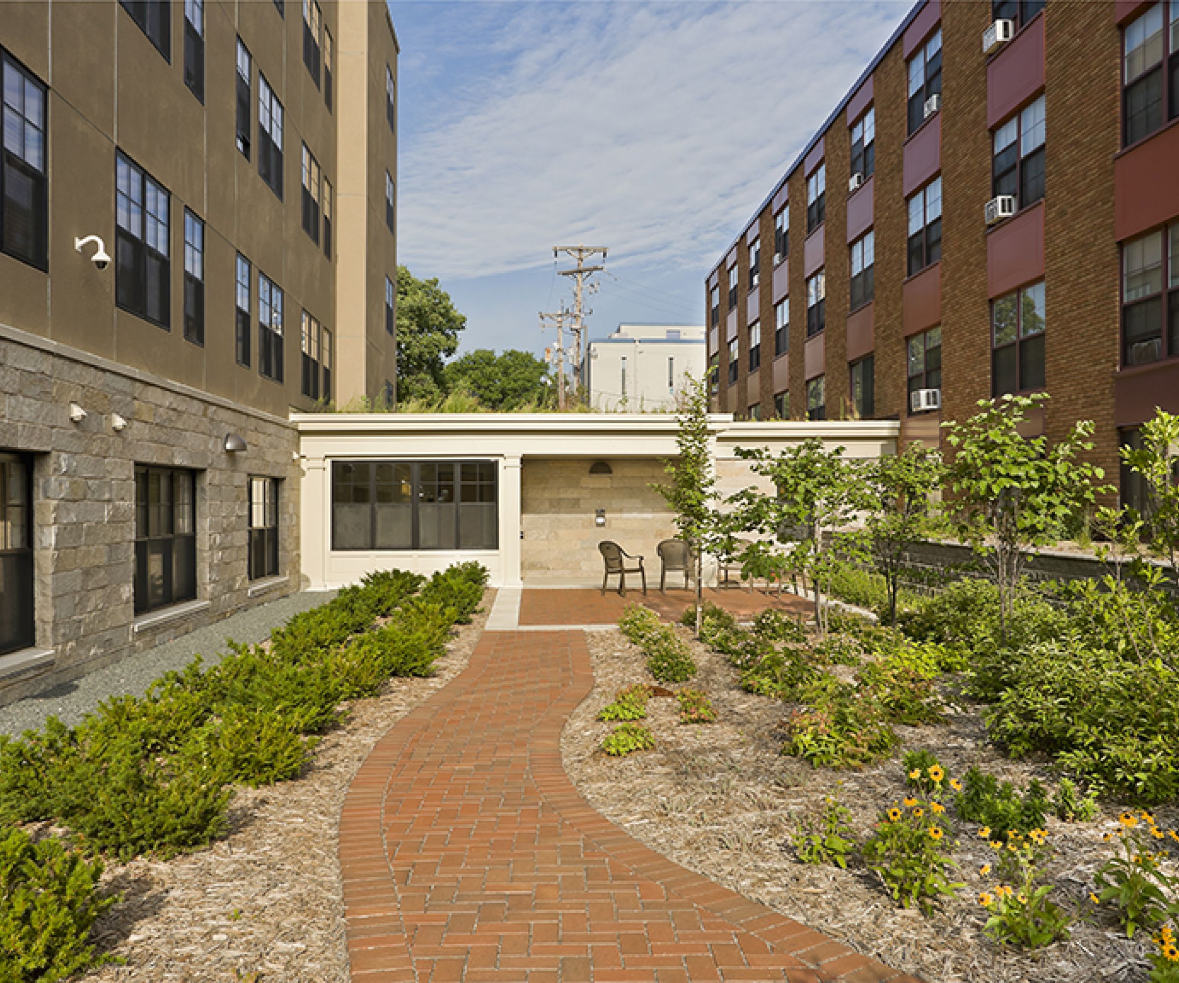 Aeon Affordable Homes and Community A brick path through a garden courtyard, surrounded by affordable apartment homes with large windows and bright flowers.