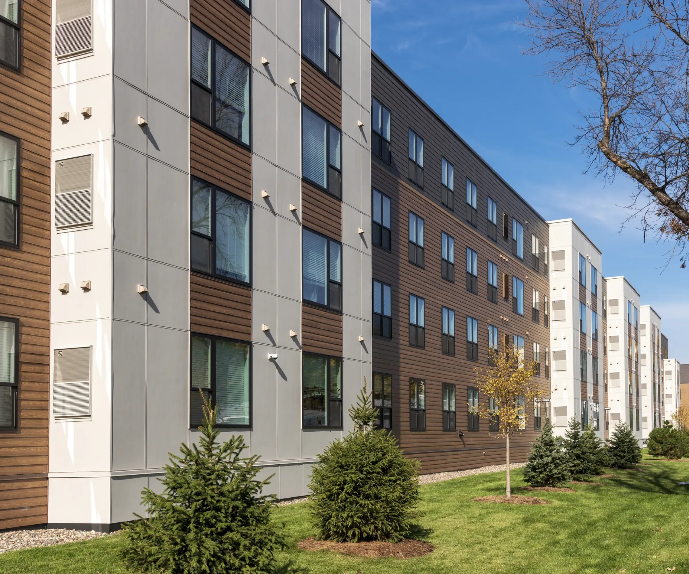 Aeon Affordable Homes and Community Affordable modern apartment homes with a wood and white panel facade, surrounded by trees and grass under a clear blue sky.