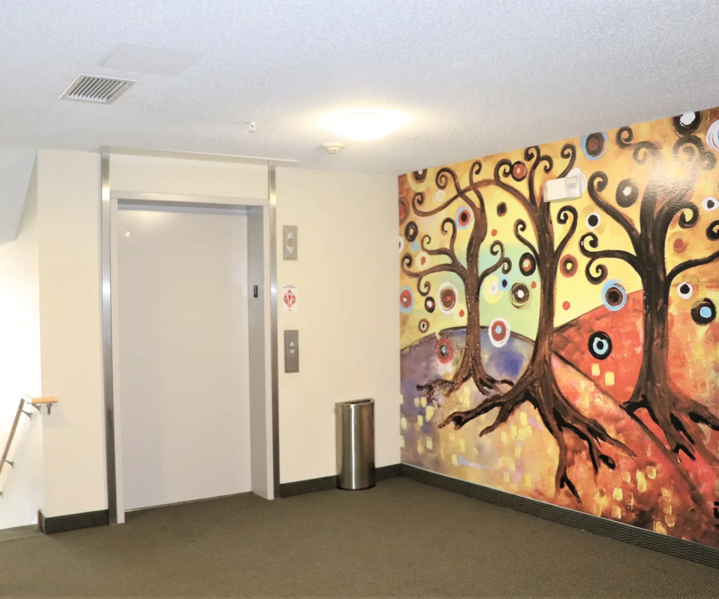 Aeon Affordable Homes and Community Colorful tree mural adorns the elevator entrance of these affordable apartment homes, with a small trash can nearby.