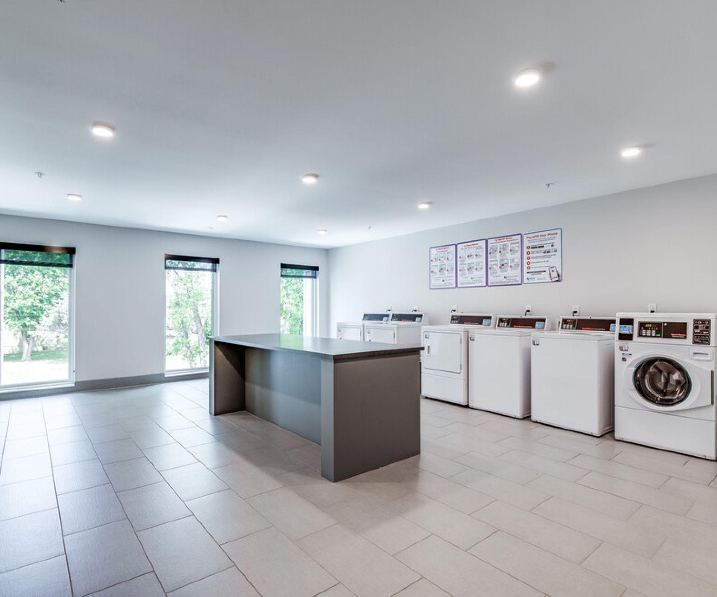 Aeon Affordable Homes and Community Laundry room in affordable apartment homes, featuring several washing machines, a central folding table, large windows, and bright lighting.