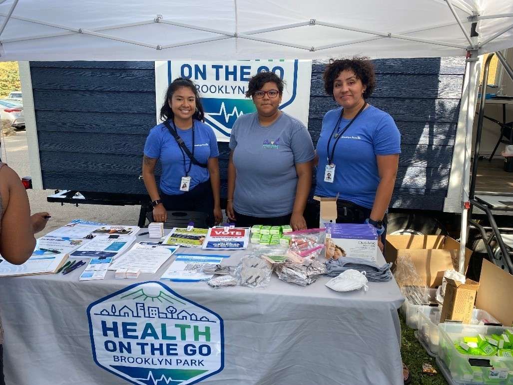 Aeon Affordable Homes and Community Three people at a community health booth with pamphlets and health supplies on a table under a canopy. Aeon Affordable Homes and Community Three people in blue shirts stand behind a table with health flyers and supplies under a canopy. Health on the Go tabling Health on the Go tabling