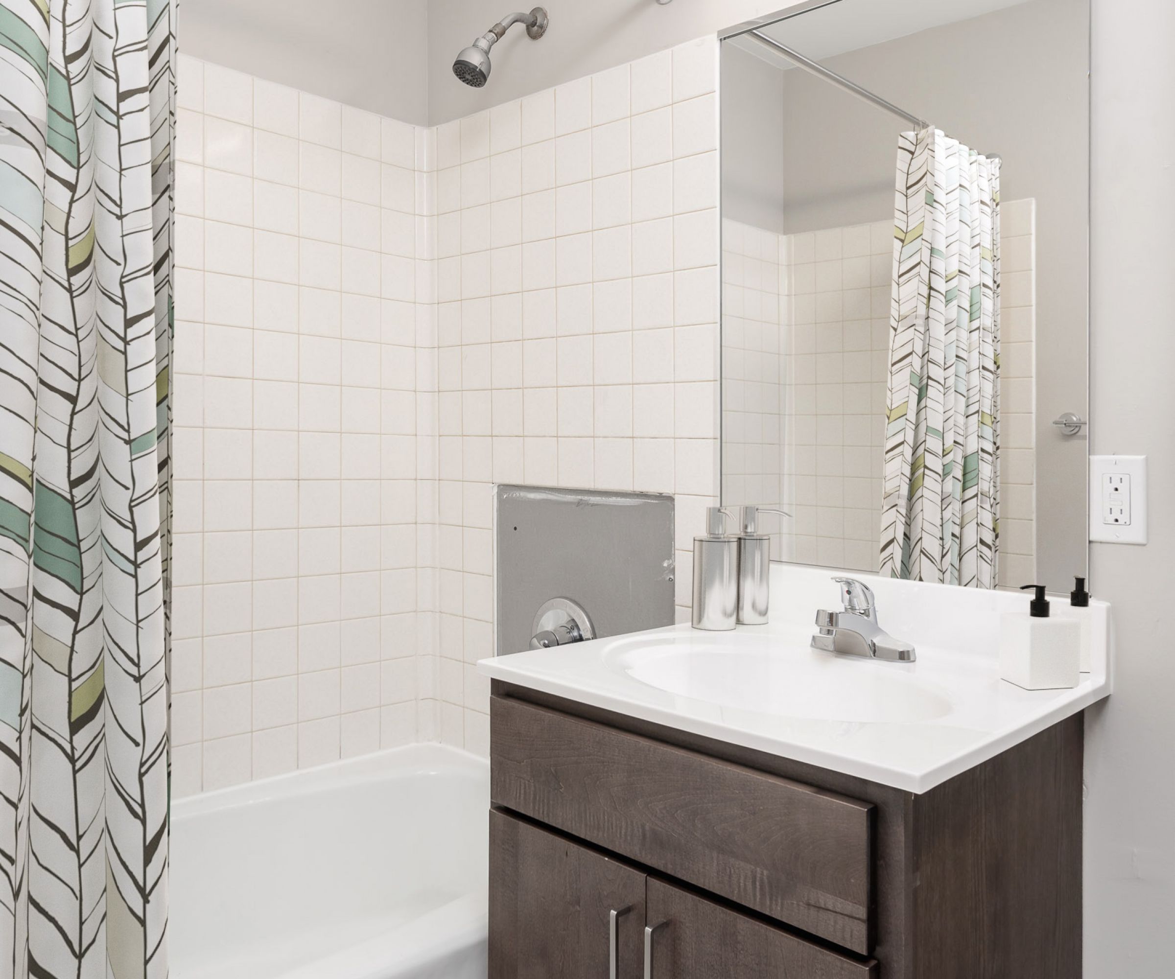 Aeon Affordable Homes and Community Modern bathroom in affordable apartment homes featuring a shower-tub combo, patterned curtain, mirror above dark wood vanity, and wall sconce light.
