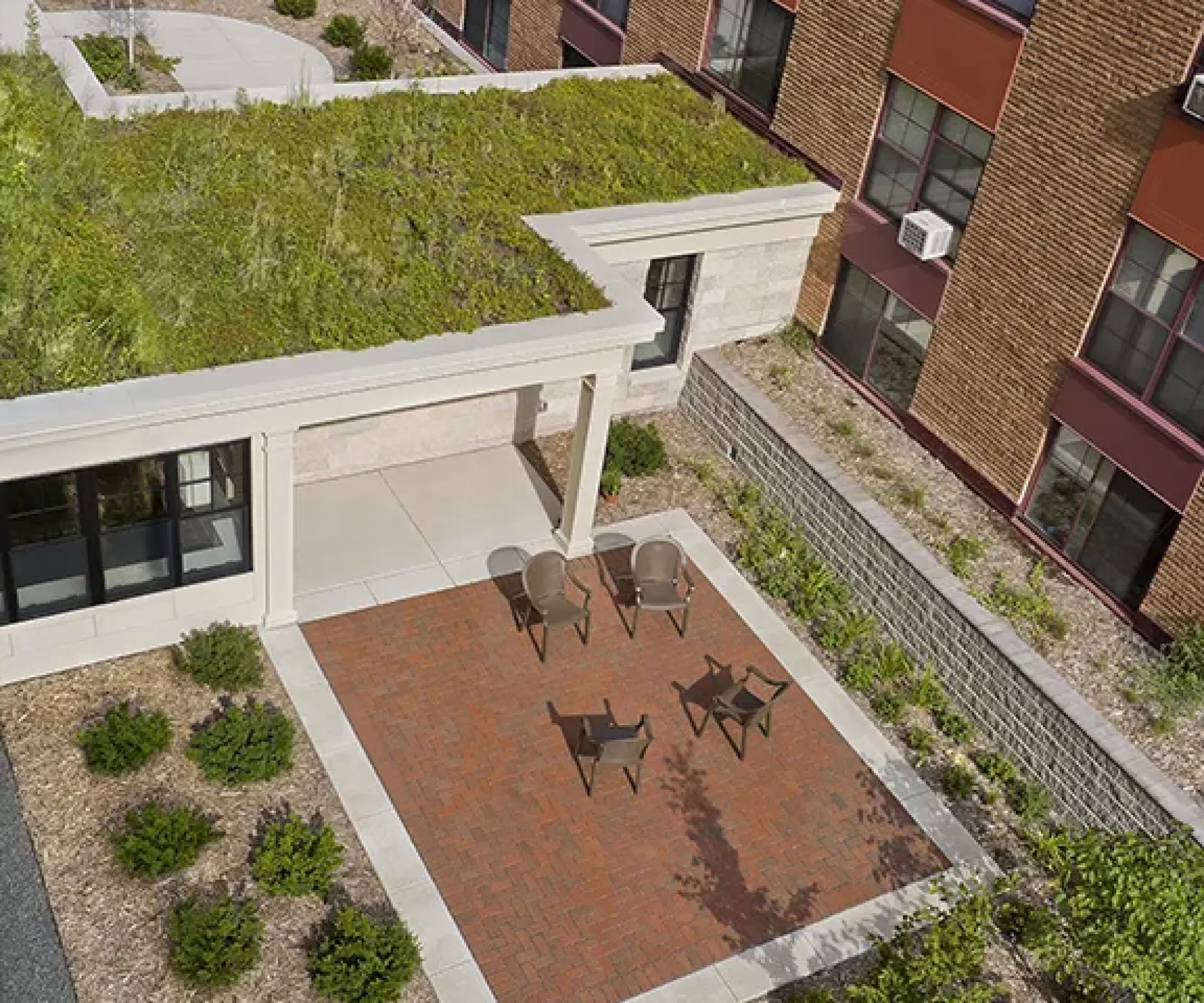 Aeon Affordable Homes and Community Aerial view of a roof garden on affordable apartment homes with brick patio, three chairs, and surrounding plants next to a brick building.