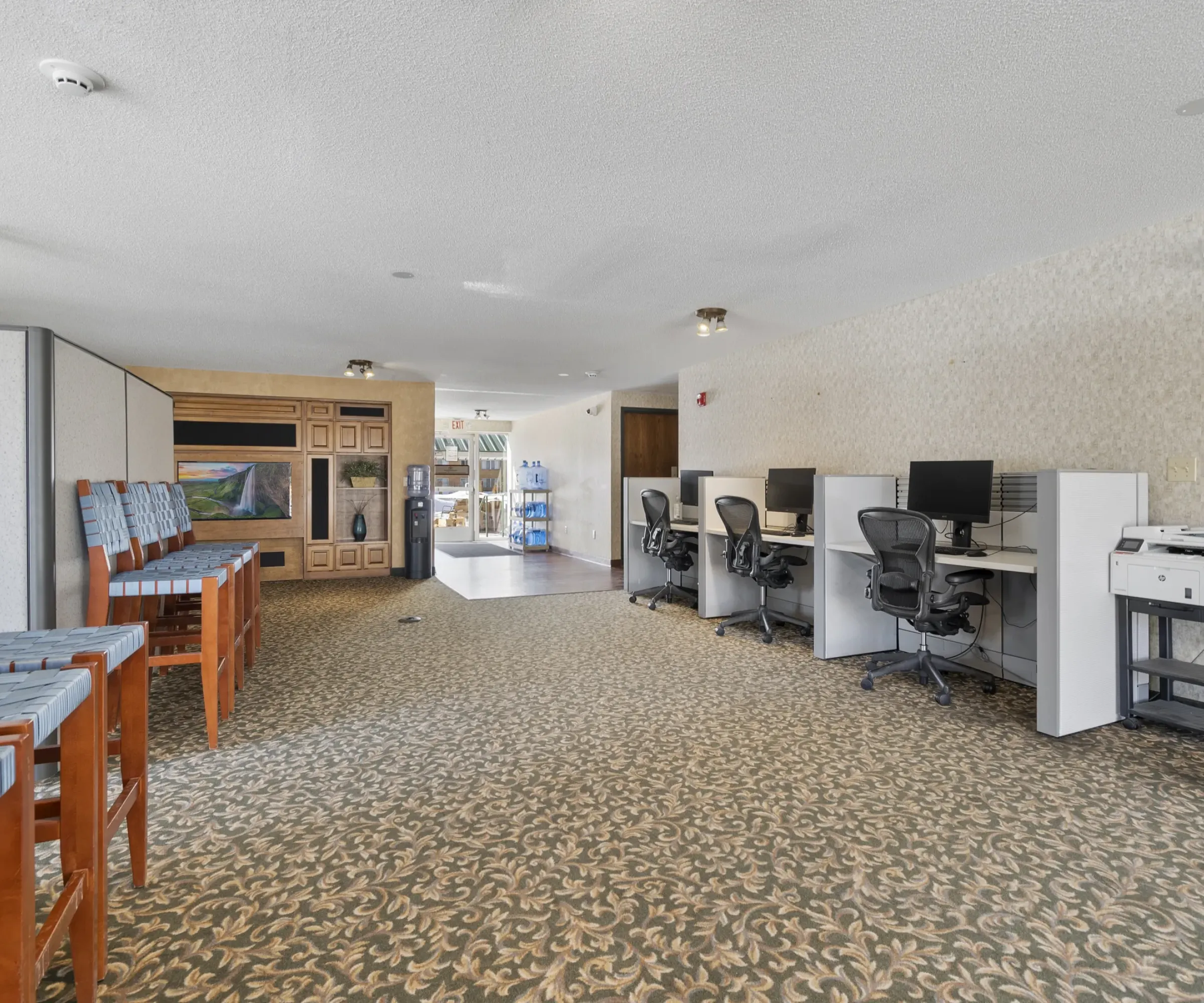 Aeon Affordable Homes Room with computer stations, chairs, and a copier on a patterned carpet. Natural light from windows in the background.