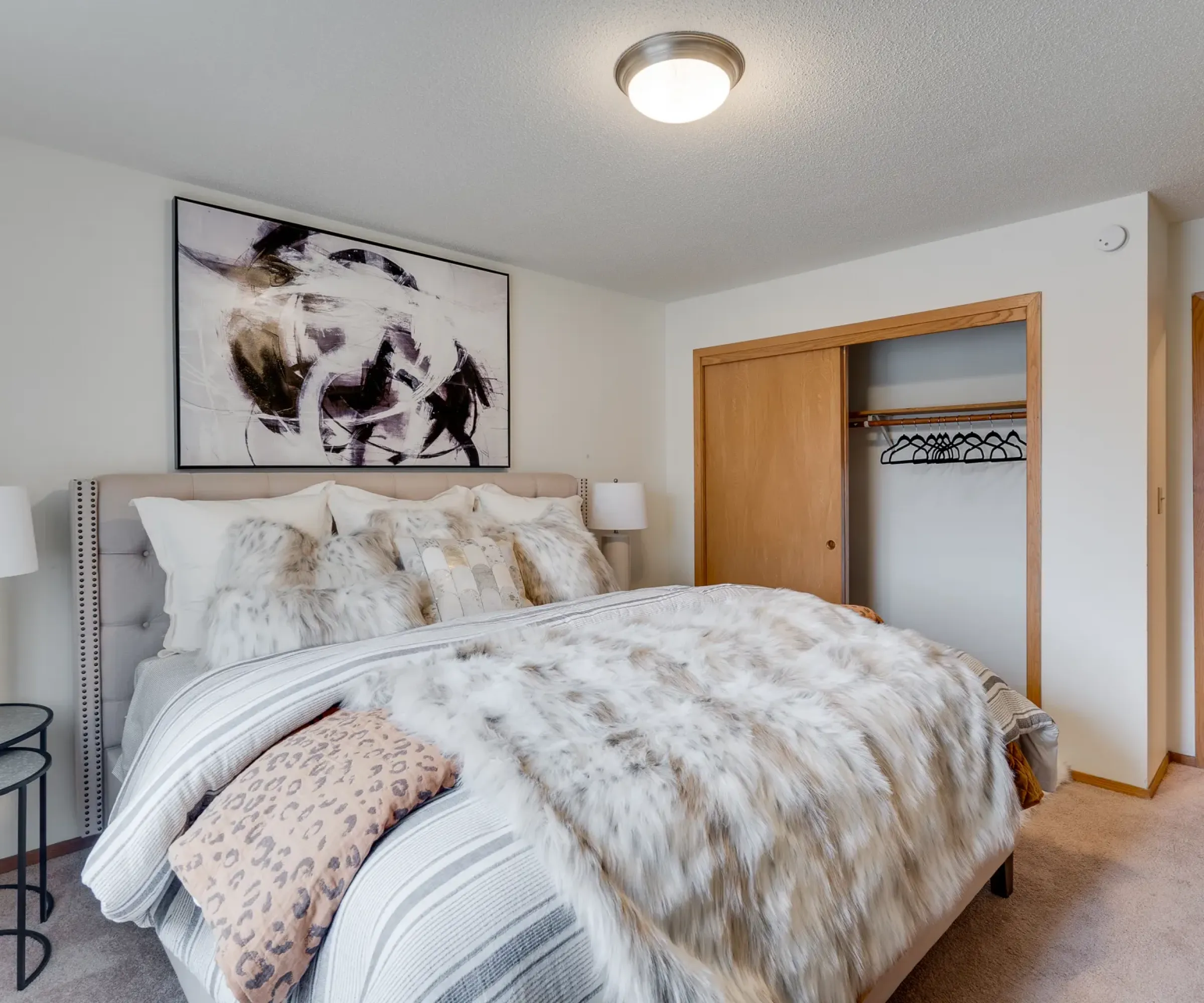 Aeon Affordable Homes Bedroom with a large bed, fur throws, abstract artwork, and wooden closet doors in a neutral-toned room.