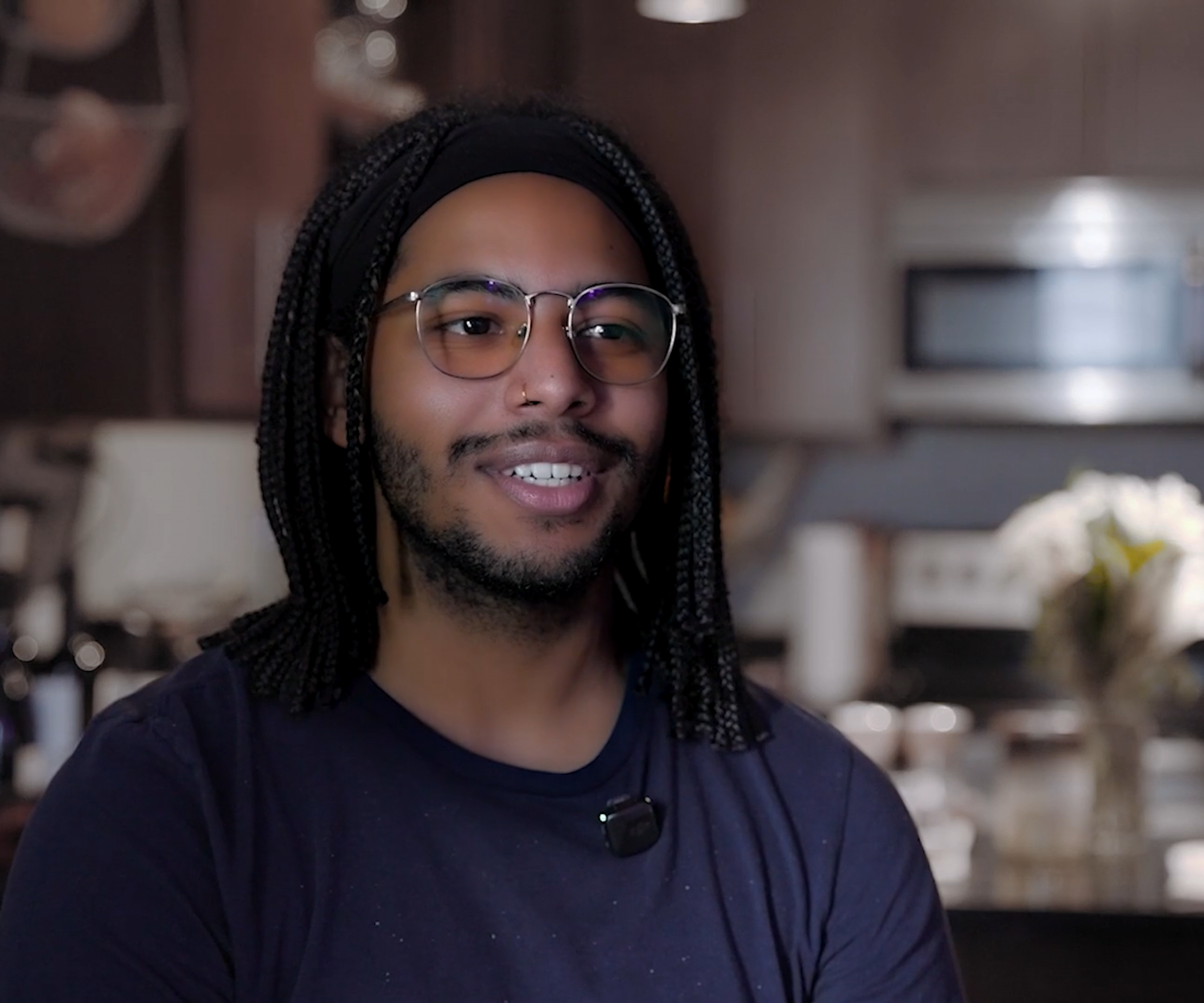 Aeon Affordable Homes A smiling person with glasses and braids sits in a kitchen, wearing a dark shirt and headset microphone.