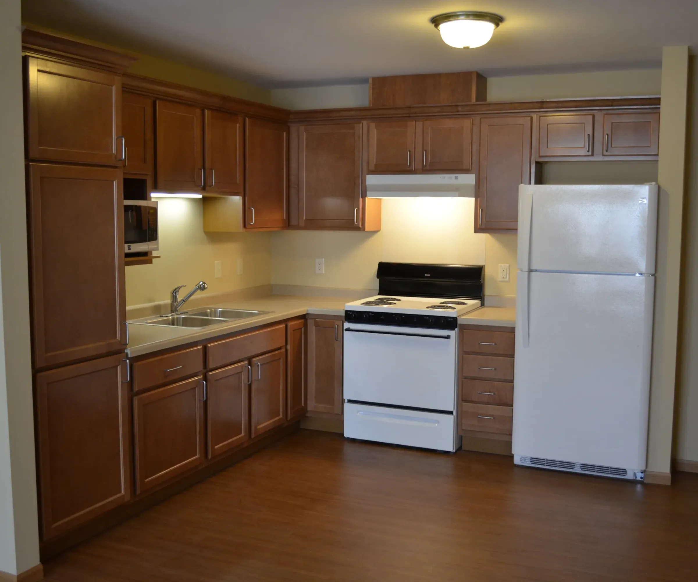 Aeon Affordable Homes and Community A modern kitchen in affordable apartment homes, featuring wooden cabinets, white appliances, and a hardwood floor.