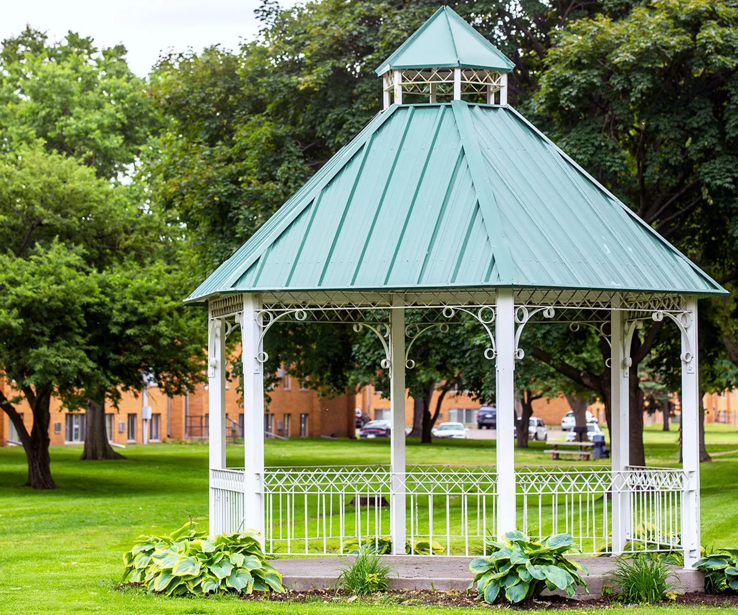 Aeon Affordable Homes A white gazebo with a green roof in a grassy park, surrounded by trees and buildings in the background.