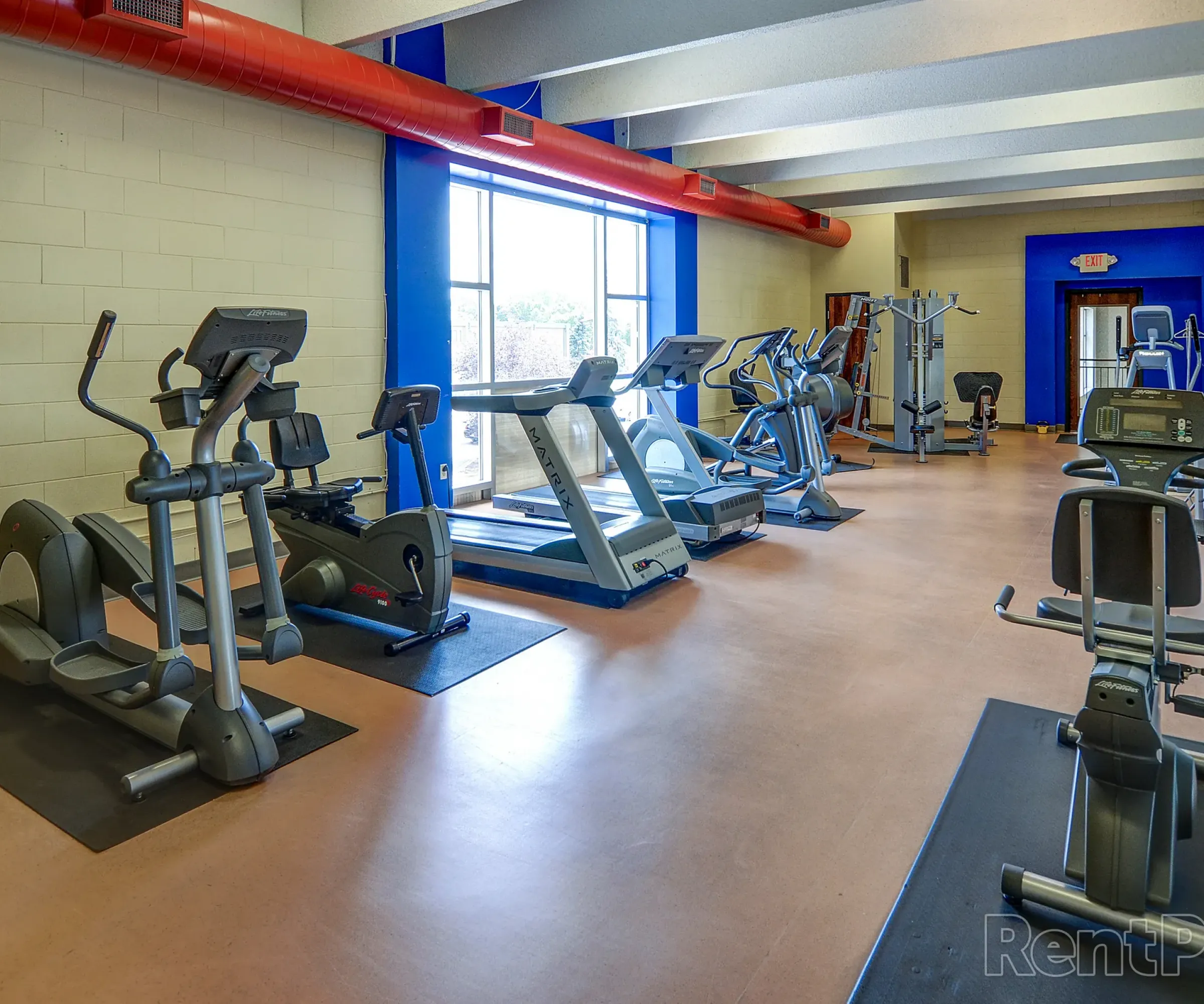 Aeon Affordable Homes Gym with various exercise machines, including treadmills and ellipticals, with large windows and a red ceiling pipe.