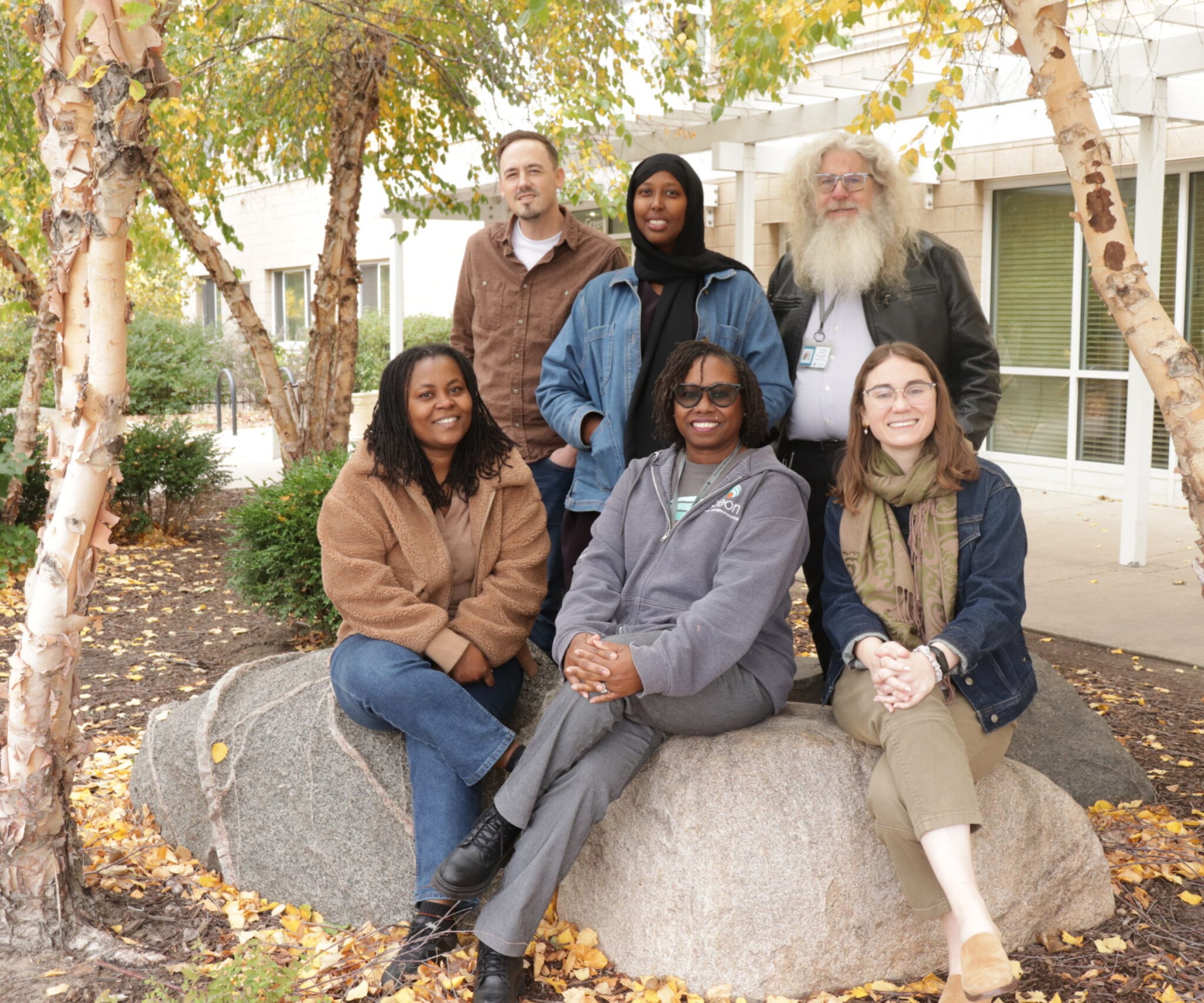 Aeon Affordable Homes Six people pose and smile outdoors in autumn, sitting and standing on large rocks among birch trees.