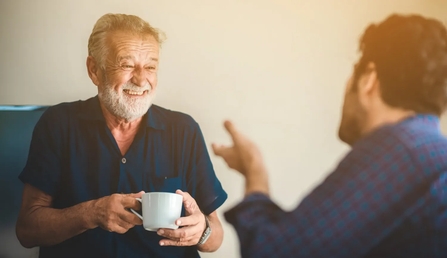 Aeon Affordable Homes with an elderly man smiling and holding a mug while talking with another person, who gestures in conversation.
