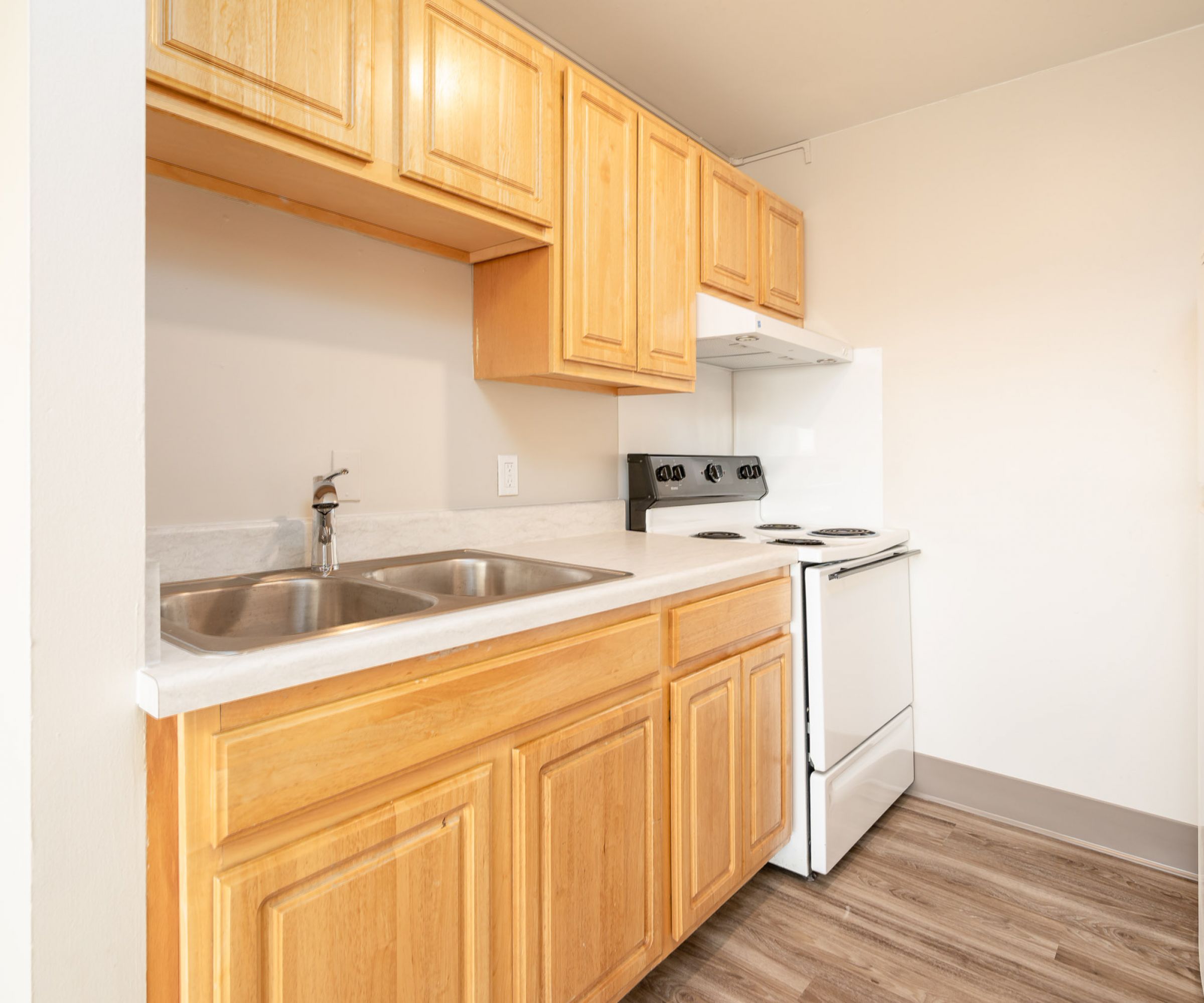Aeon Affordable Homes and Community Affordable apartment homes featuring a small kitchen with wooden cabinets, a double sink, white stove, and beige walls.