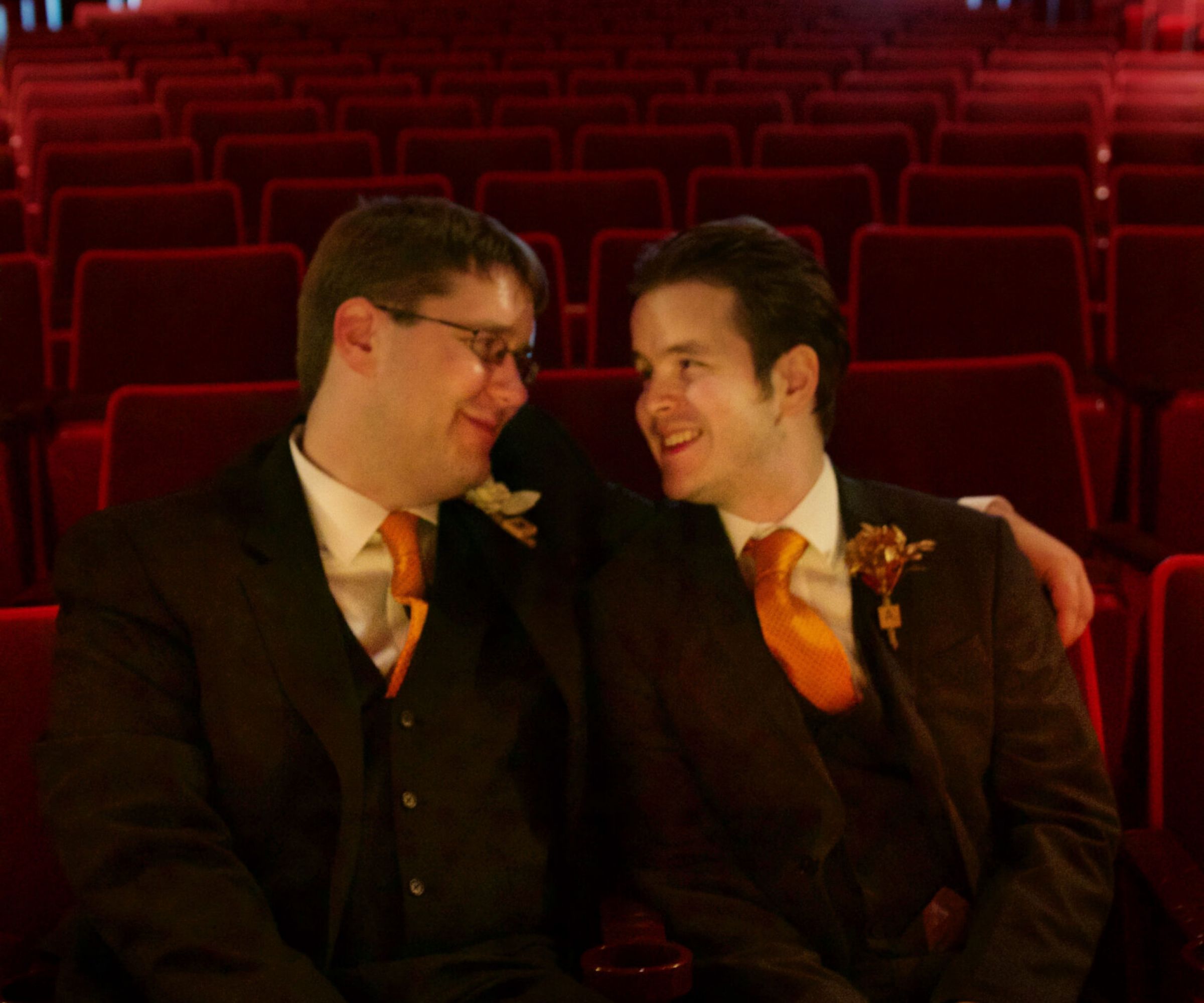 Aeon Affordable Homes Two men in suits with orange ties sit closely together, smiling, in an empty theater with red seats—it's part of Andrew's Story.