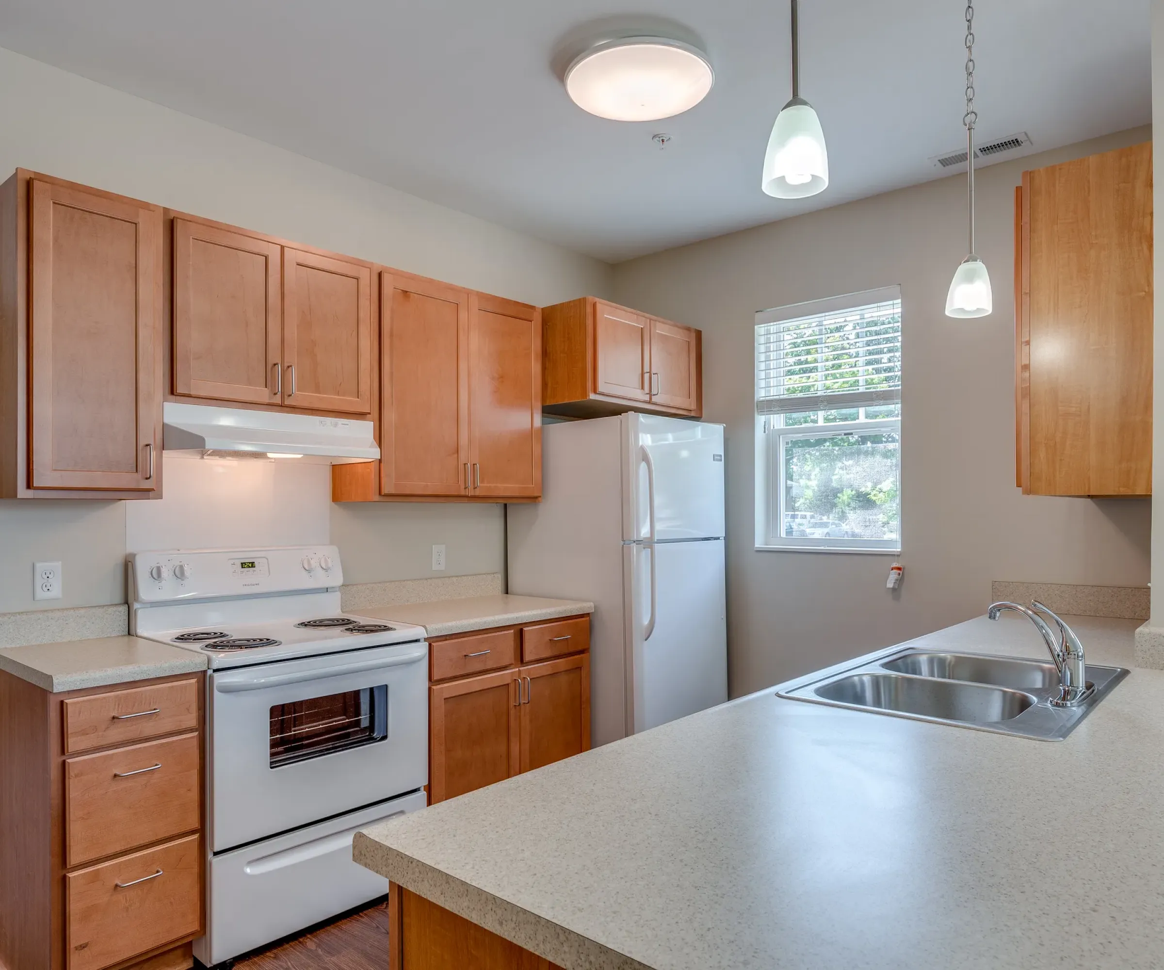 Aeon Affordable Homes and Community Modern kitchen in affordable apartment homes features wooden cabinets, white appliances, double sink, pendant lights, and bright windows.