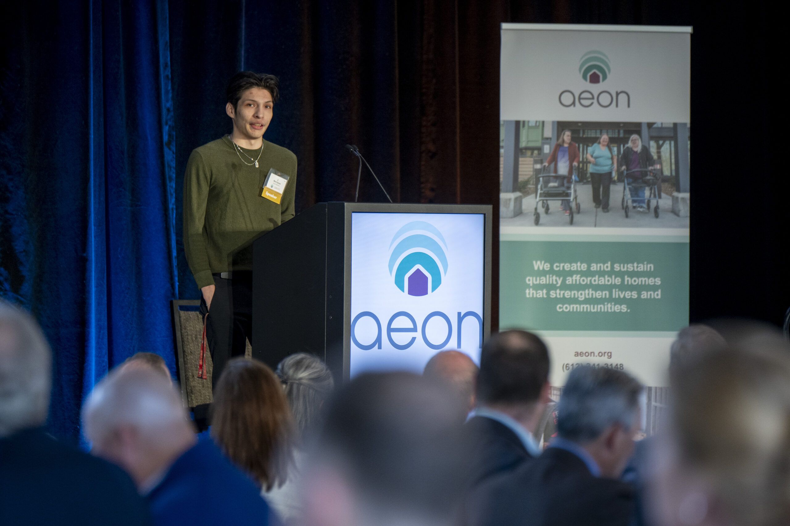 Aeon Affordable Homes A person speaks at a podium labeled "aeon" during an event, with an audience and banner visible.