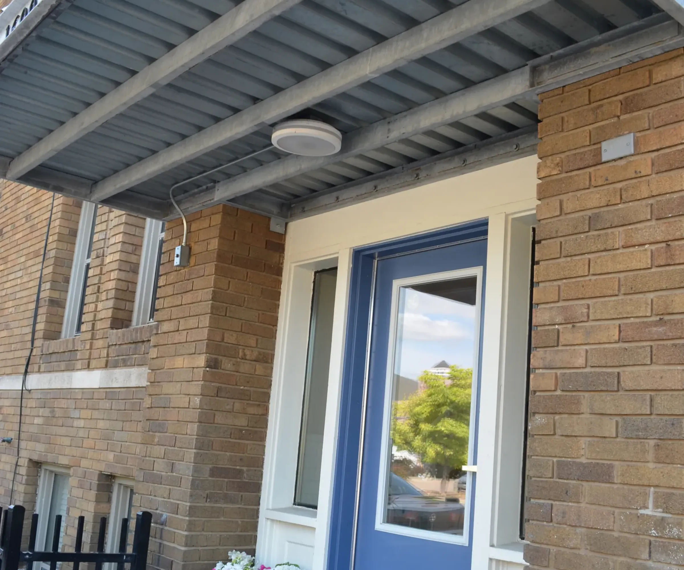 Aeon Affordable Homes and Community Blue door with glass panels, metal awning, and planters adorn these affordable apartment homes in a charming brick building.