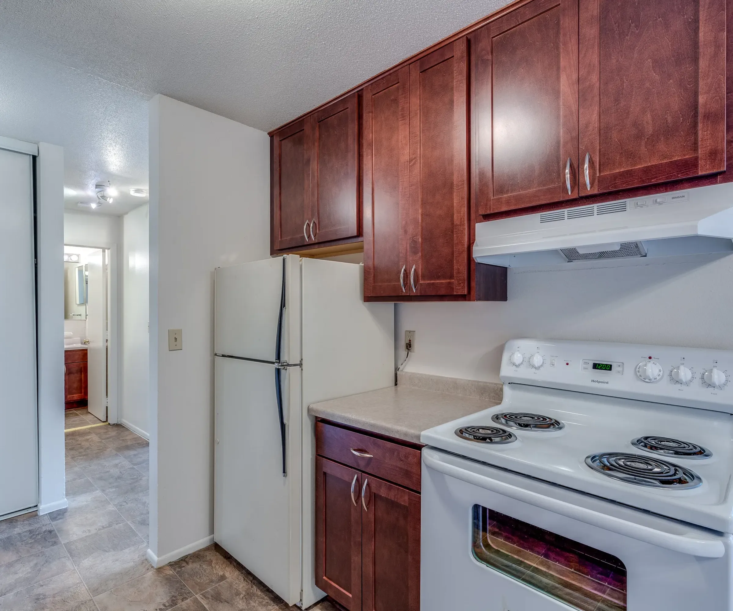 Aeon Affordable Homes and Community Small kitchen in affordable apartment home with dark wooden cabinets, white appliances, and tiled floor leading to a narrow hallway.