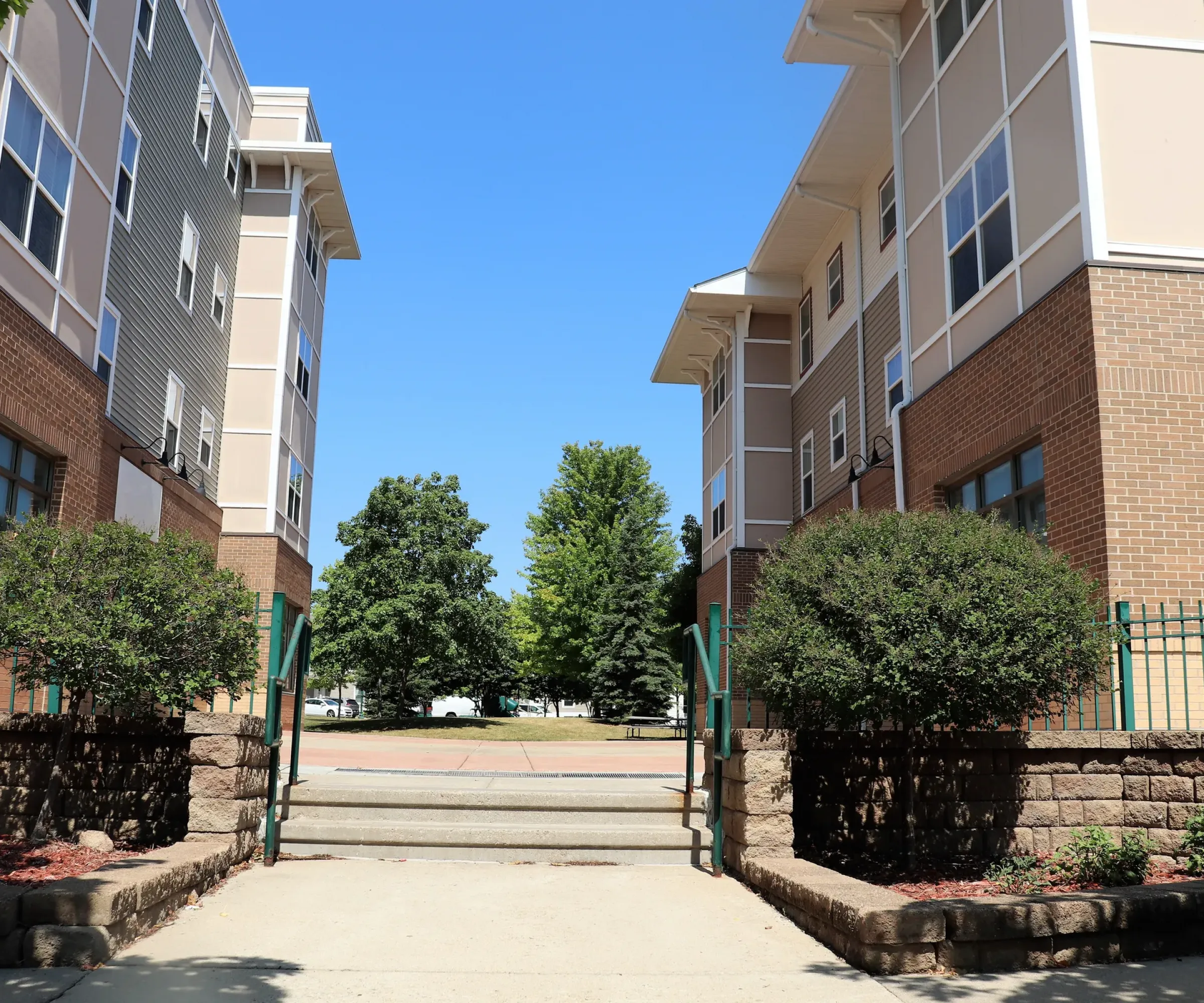 Aeon Affordable Homes and Community Entrance with green gates leads to courtyard between two modern brick buildings, offering affordable apartment homes; trees visible in background.