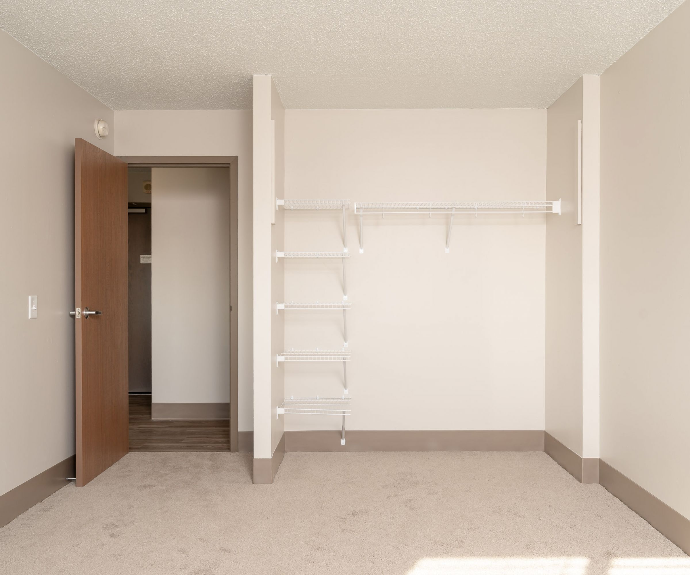 Aeon Affordable Homes and Community Affordable apartment homes featuring an empty room with beige carpet, an open wooden door, and white wall shelves.
