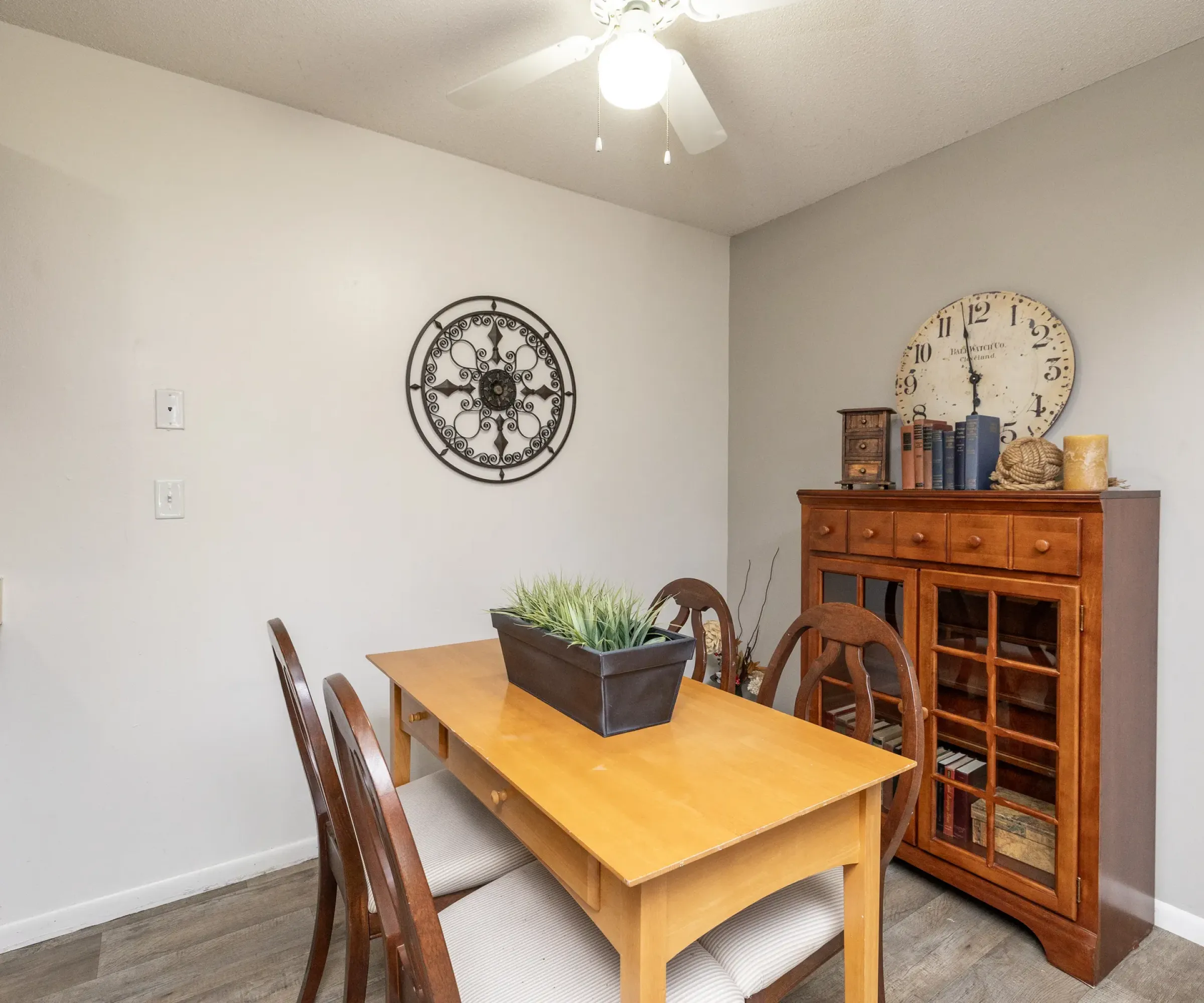 Aeon Affordable Homes Dining area in Blooming Meadows South: wooden table, chairs, wall clock, cabinet with books and decor. Ceiling fan overhead.