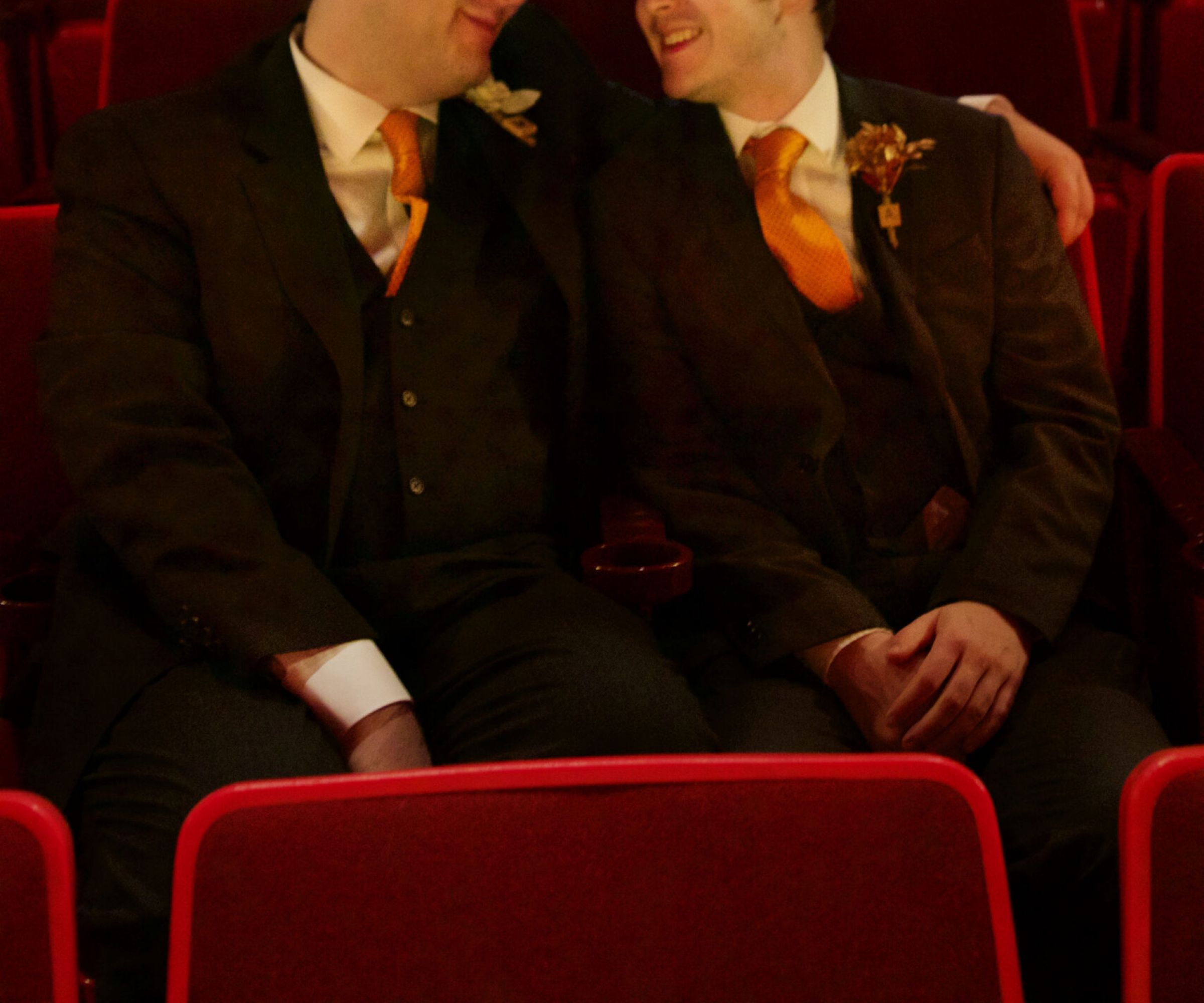 Aeon Affordable Homes Two men in suits with orange ties sit closely together, smiling, in an empty theater with red seats—it's part of Andrew's Story.