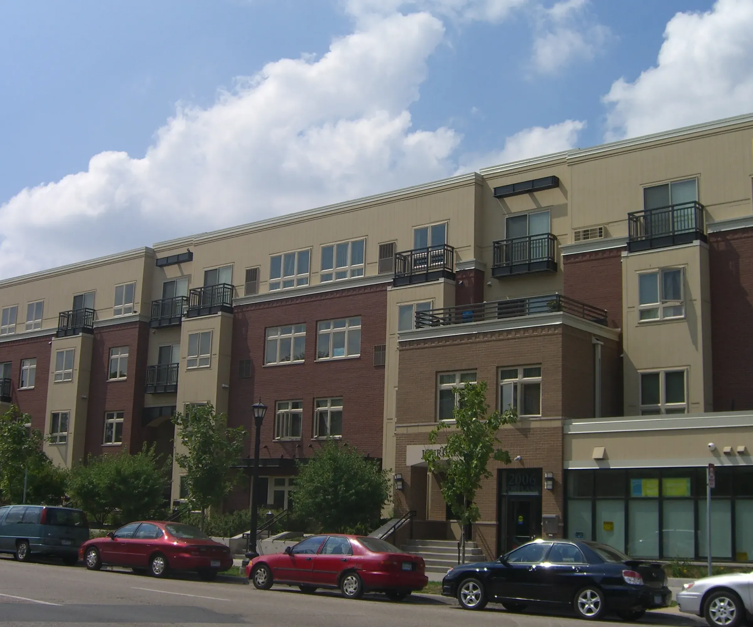 Aeon Affordable Homes and Community Affordable apartment homes in a four-story building with red brick, surrounded by trees and parked cars on a sunny day.