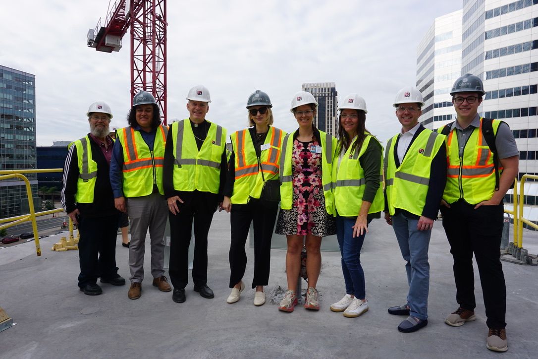 Aeon Affordable Homes Eight people in hard hats and safety vests stand together on a rooftop construction site, city buildings in background.