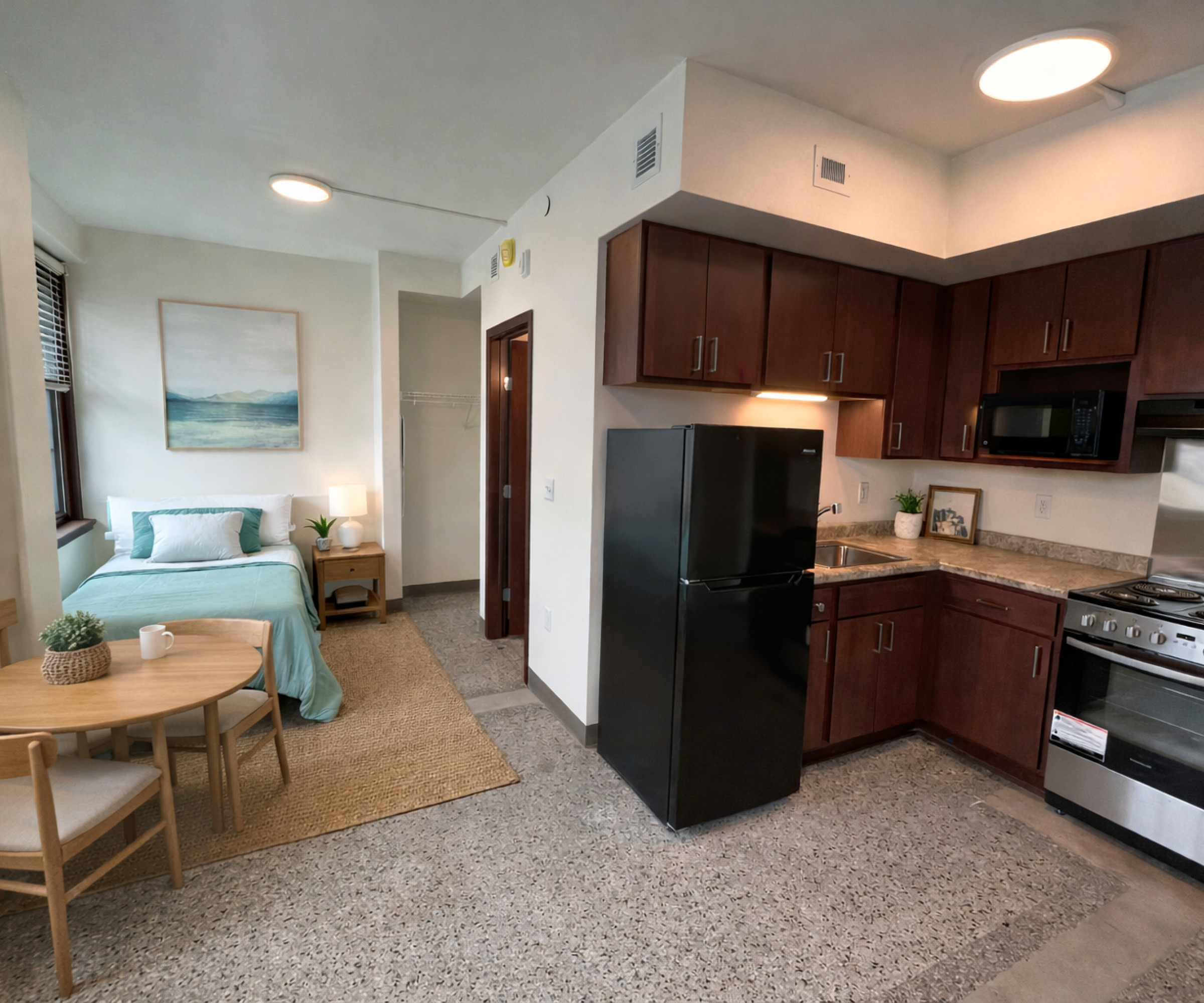 Aeon Affordable Homes Studio apartment with a kitchen, dining table, bed, and natural light from windows.