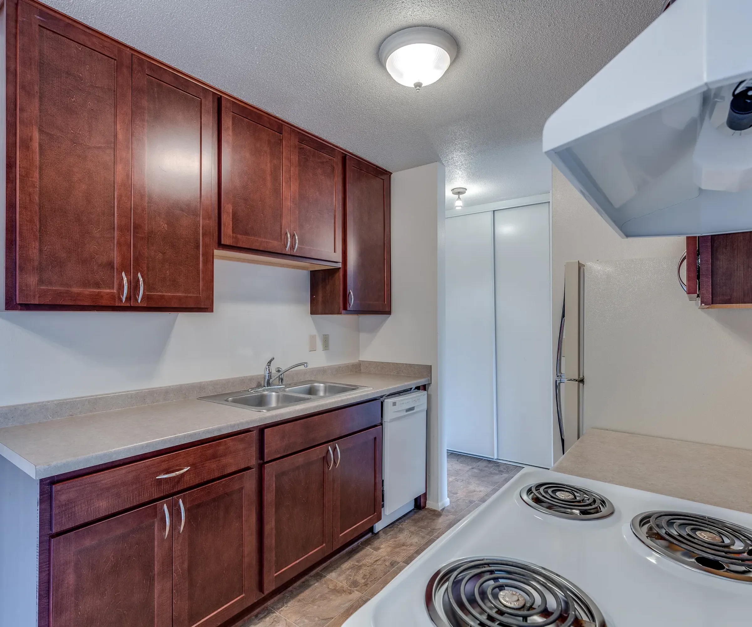 Aeon Affordable Homes and Community Compact kitchen in affordable apartment homes with dark wooden cabinets, sink, dishwasher, electric stove, and fridge under ceiling light.