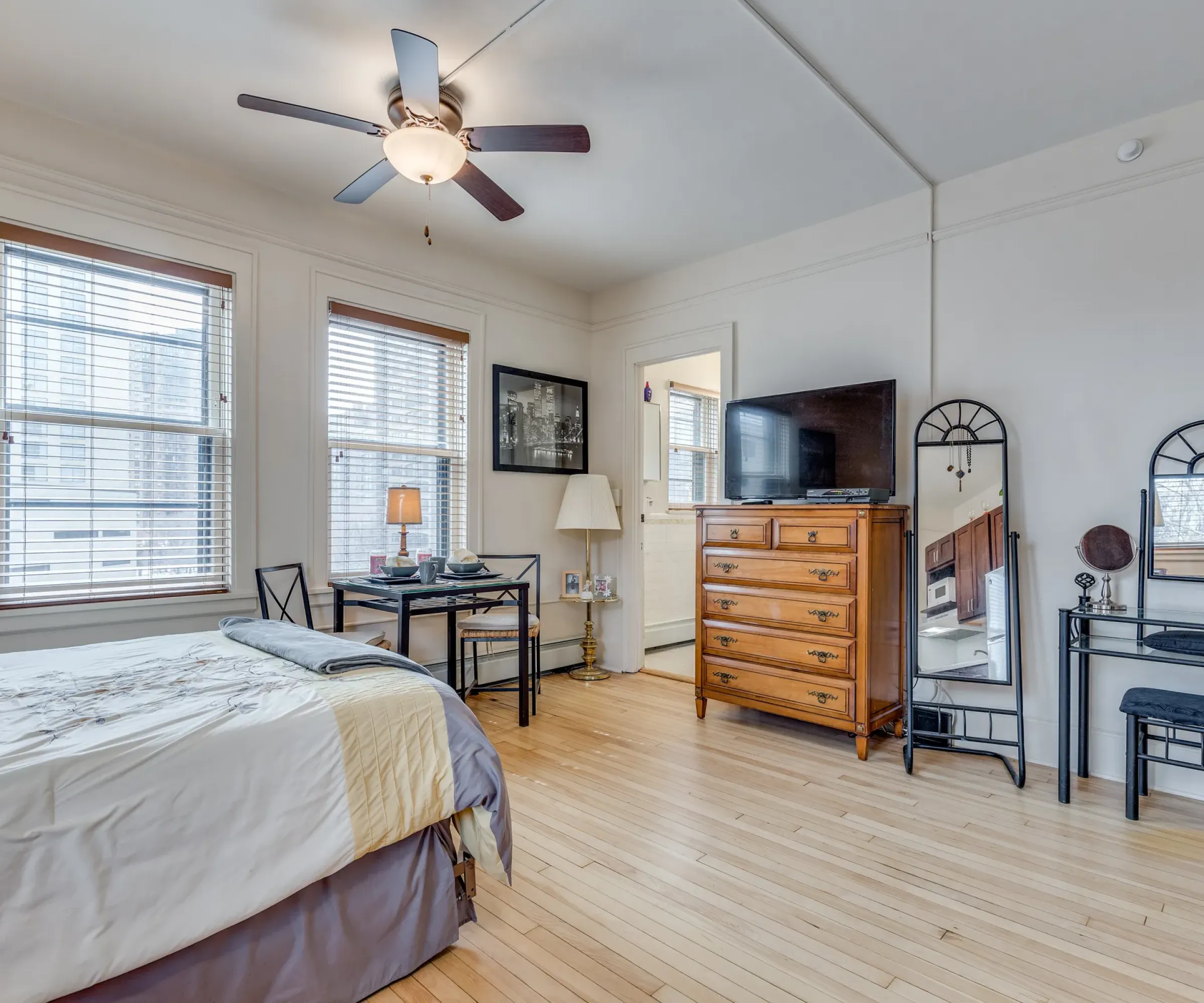 Aeon Affordable Homes and Community A cozy bedroom in affordable apartment homes with a bed, wooden dresser, TV, mirror, and ceiling fan, featuring hardwood floors and large windows.