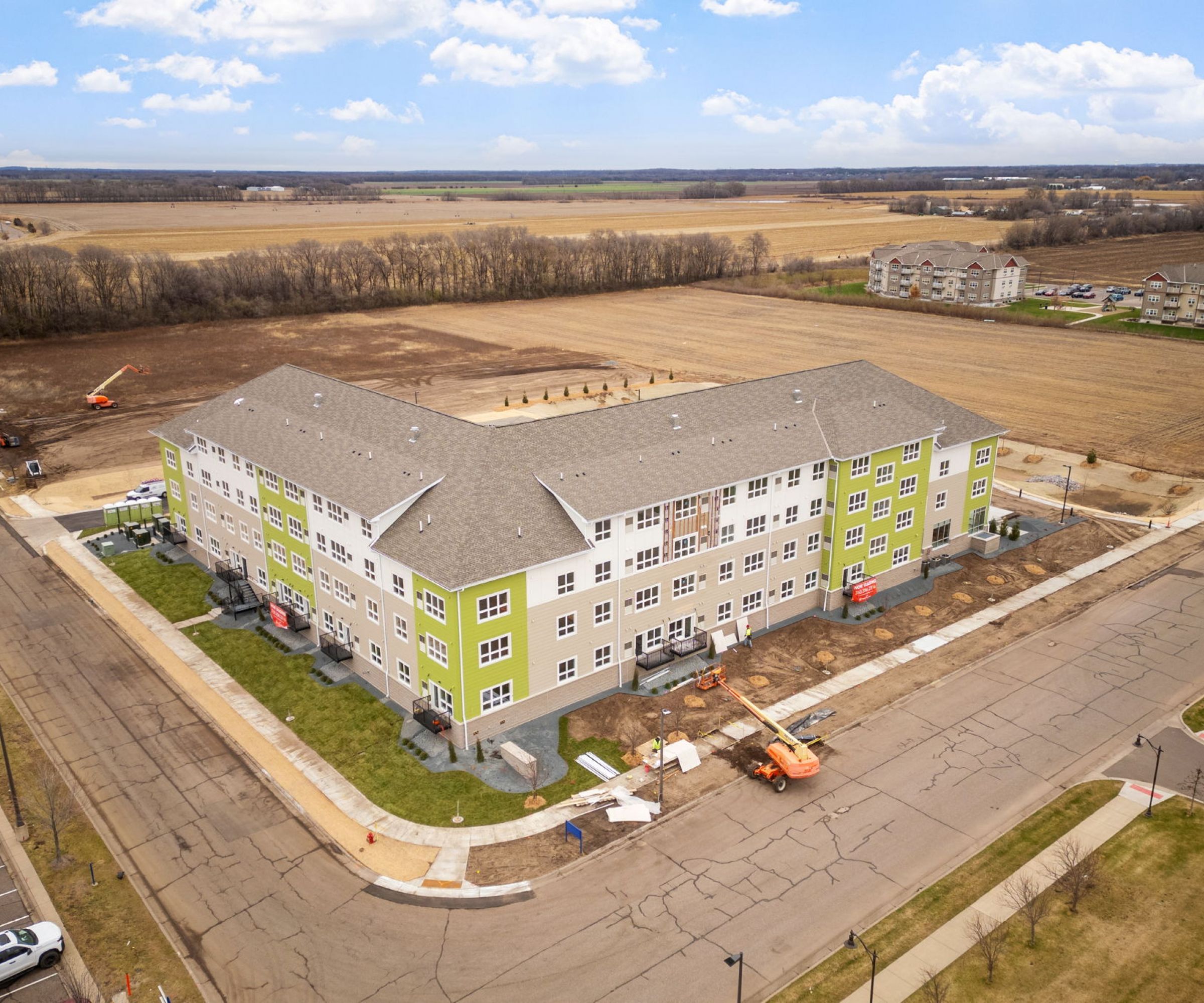 Aeon Affordable Homes and Community Aerial view of affordable apartment homes in a large L-shaped building with green accents, surrounded by construction and open fields.