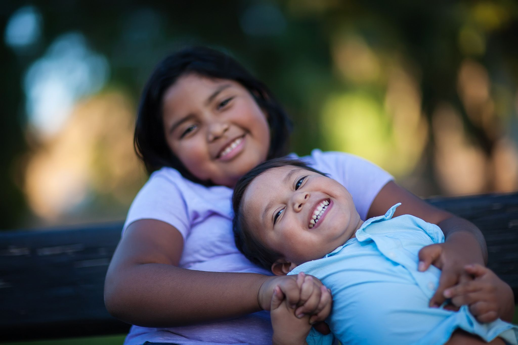 Aeon Affordable Homes Two smiling children sit close together on a bench, hugging and laughing outdoors on a sunny day.