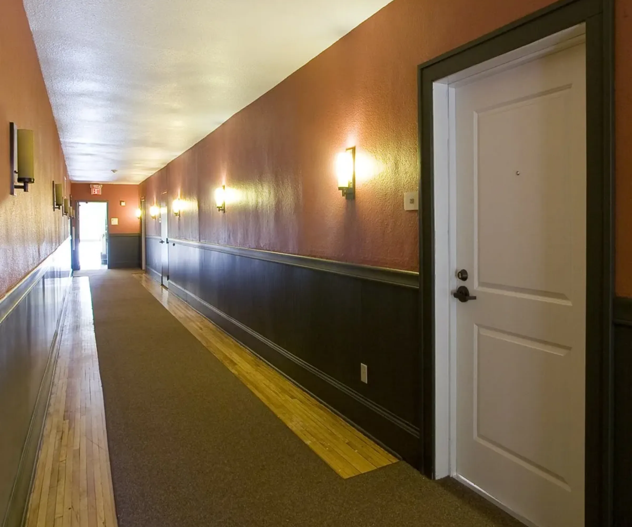 Aeon Affordable Homes Long hallway with red and brown walls, lit by wall sconces, and a white door on the right.