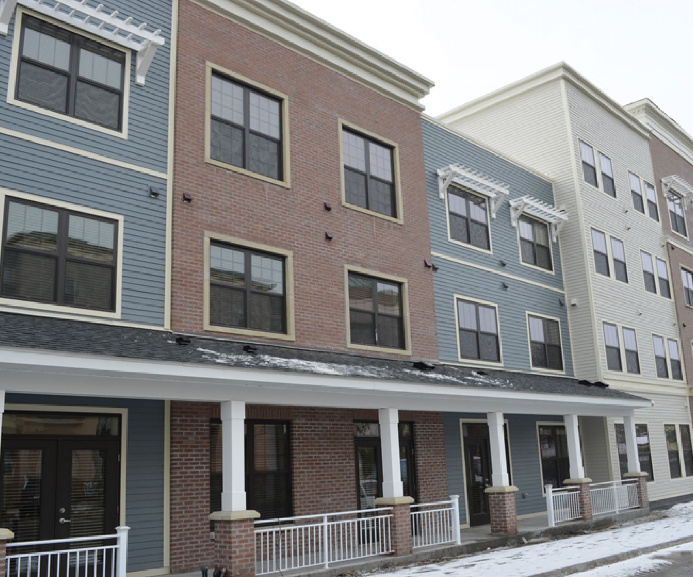 Aeon Affordable Homes and Community Three-story building with blue, brick, and beige siding, featuring balconies, snow-covered ground, and affordable apartment homes.