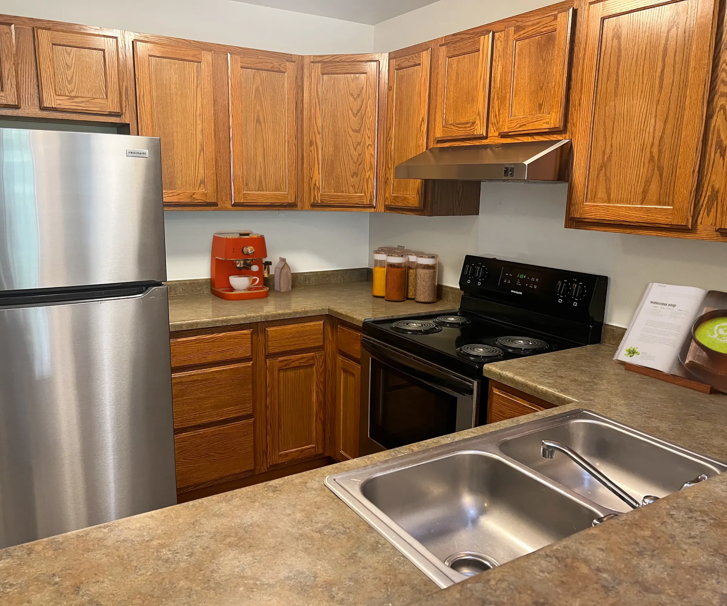 Aeon Affordable Homes and Community A kitchen in affordable apartment homes with wooden cabinets, stainless steel appliances, dual sink, and a vibrant orange coffee maker.