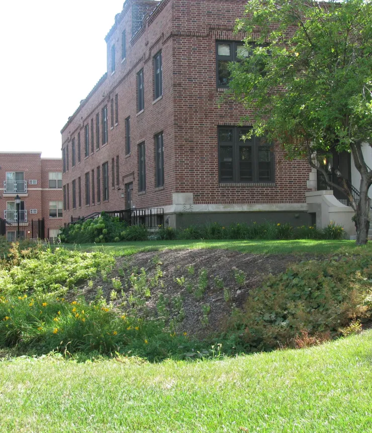 Aeon Affordable Homes and Community Affordable apartment homes in a brick building with a sloped green garden, featuring flowers, shrubs, and a tree near the entrance.