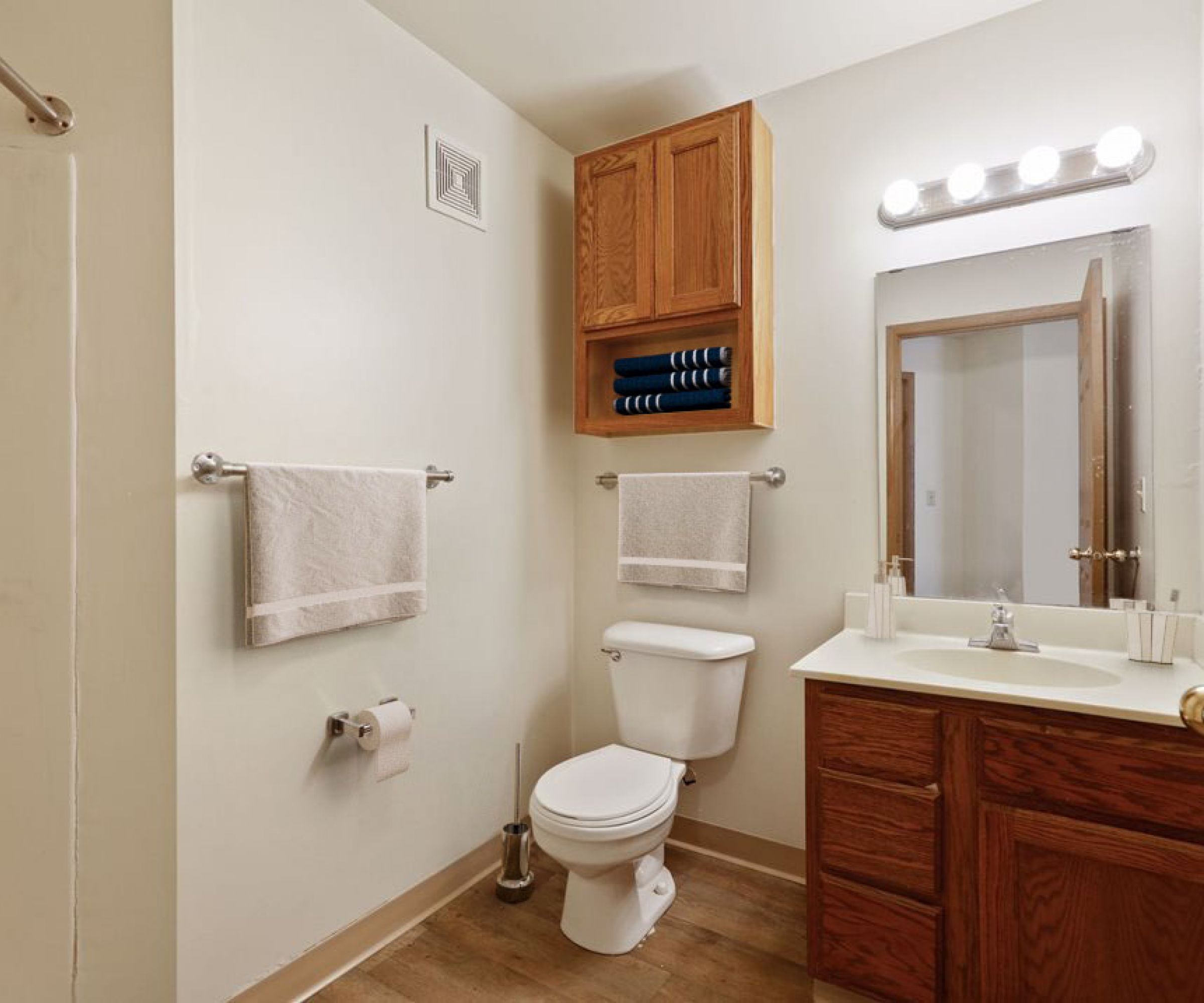 Aeon Affordable Homes and Community Affordable apartment homes feature a bathroom with wooden cabinets, a sink, mirror, and toilet. Towels hang on racks; a roll of toilet paper is on the wall.