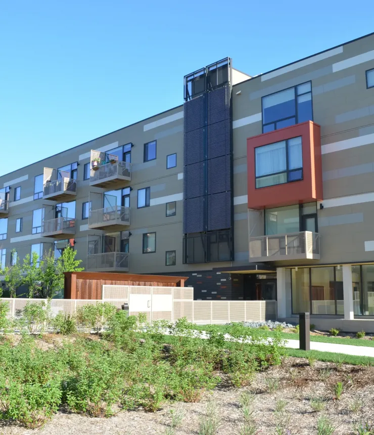 Aeon Affordable Homes and Community Modern, affordable apartment homes with balconies in a four-story building, surrounded by landscaped greenery under a clear blue sky.