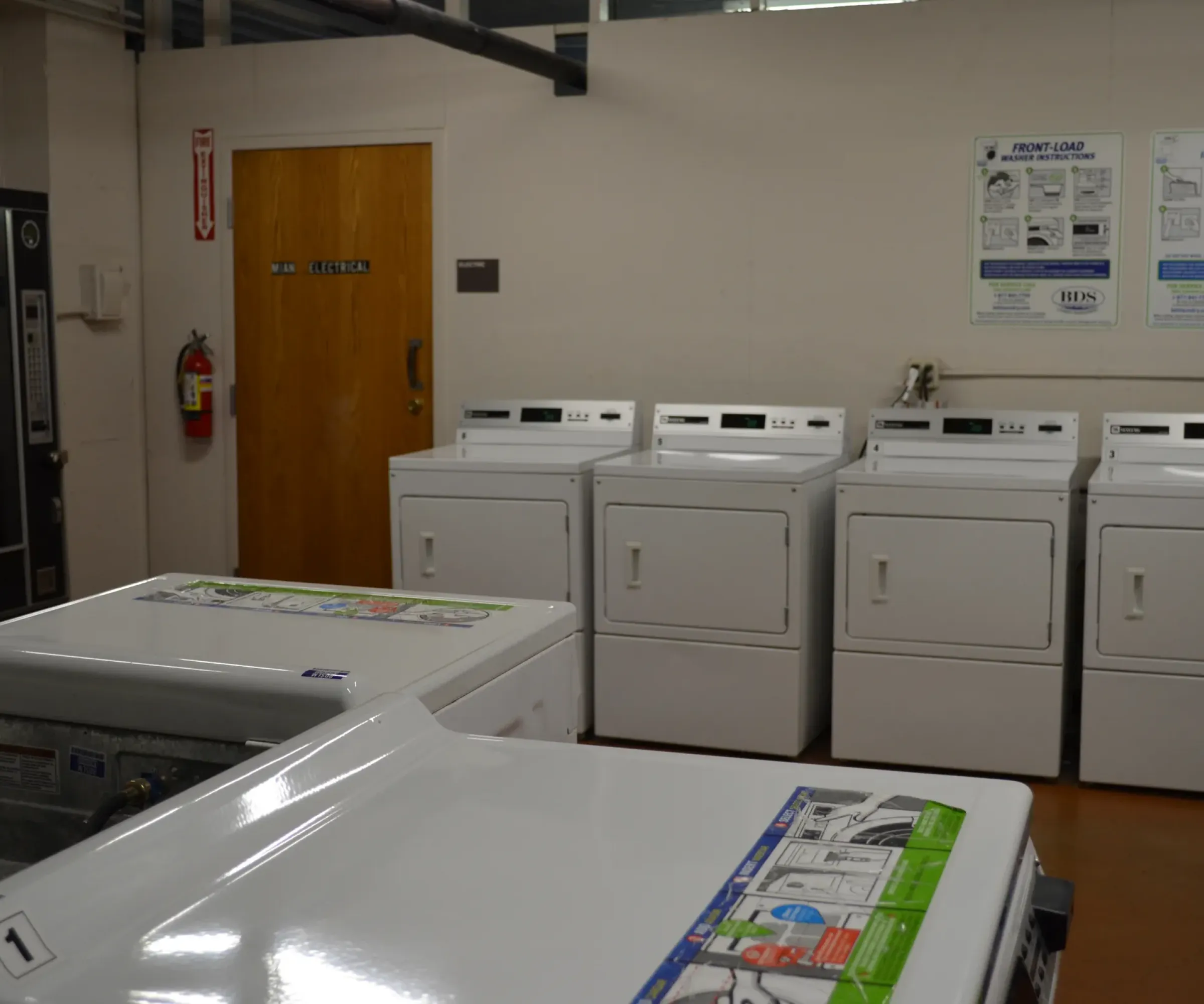 Aeon Affordable Homes and Community Laundry room in affordable apartment homes with white washers, dryers, a wooden door, and wall-mounted instructions.